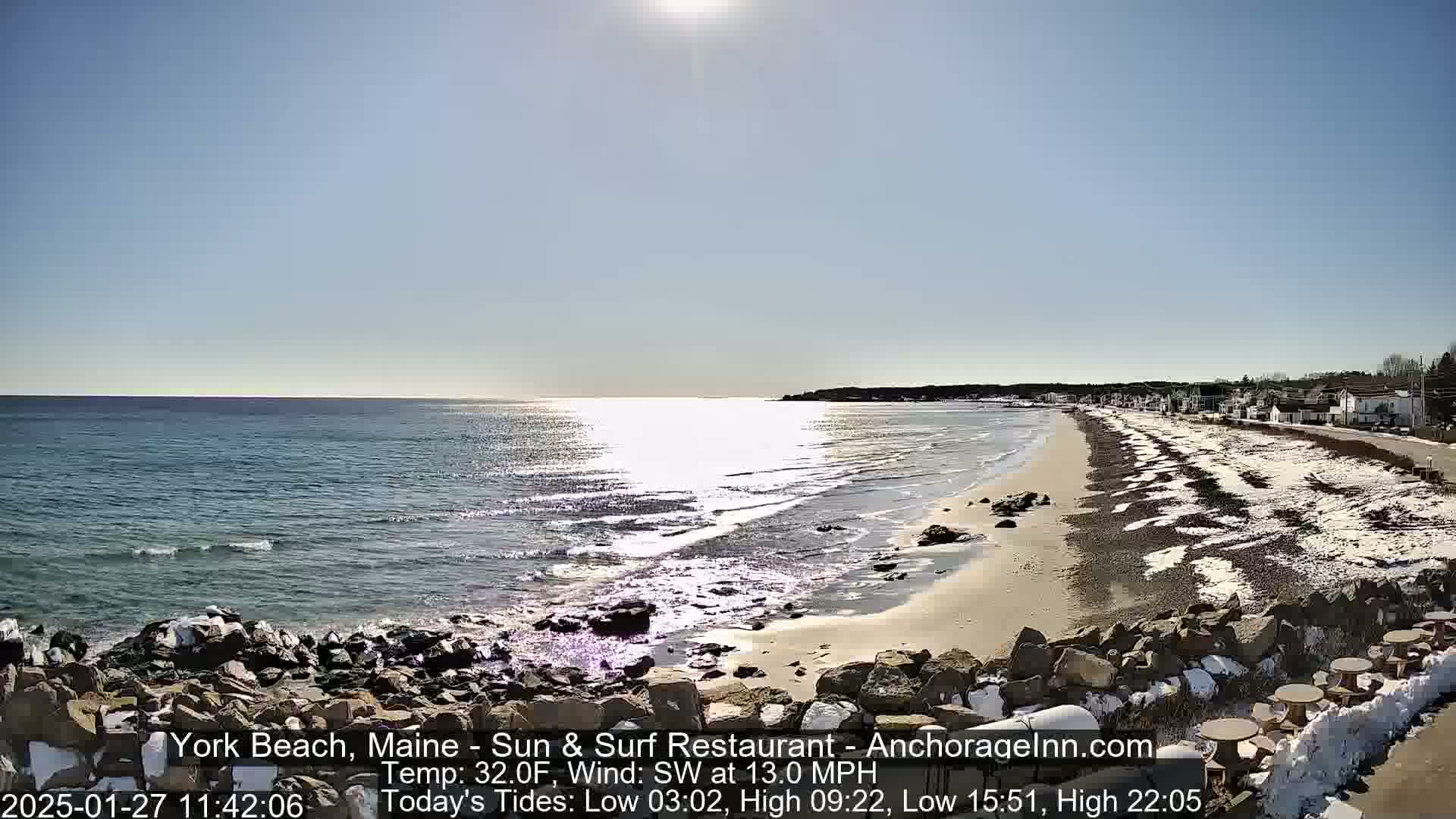 A sunny, but cold, day at a beach with a sandy shore partially covered in snow, rocky outcrops, and buildings in the distance.