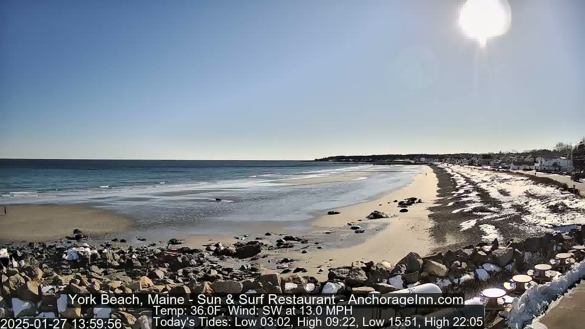 A sunny, partly snowy beach scene shows a calm ocean meeting a sandy shore with a rocky embankment and some picnic tables in the foreground.