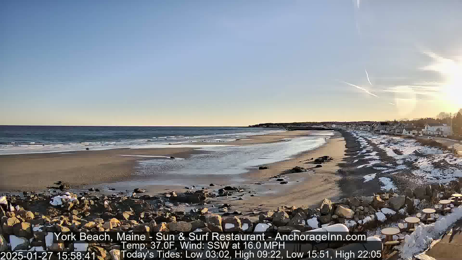 A partly sunny, cold day at a beach with snow patches on the sand and rocks near a shoreline with buildings.
