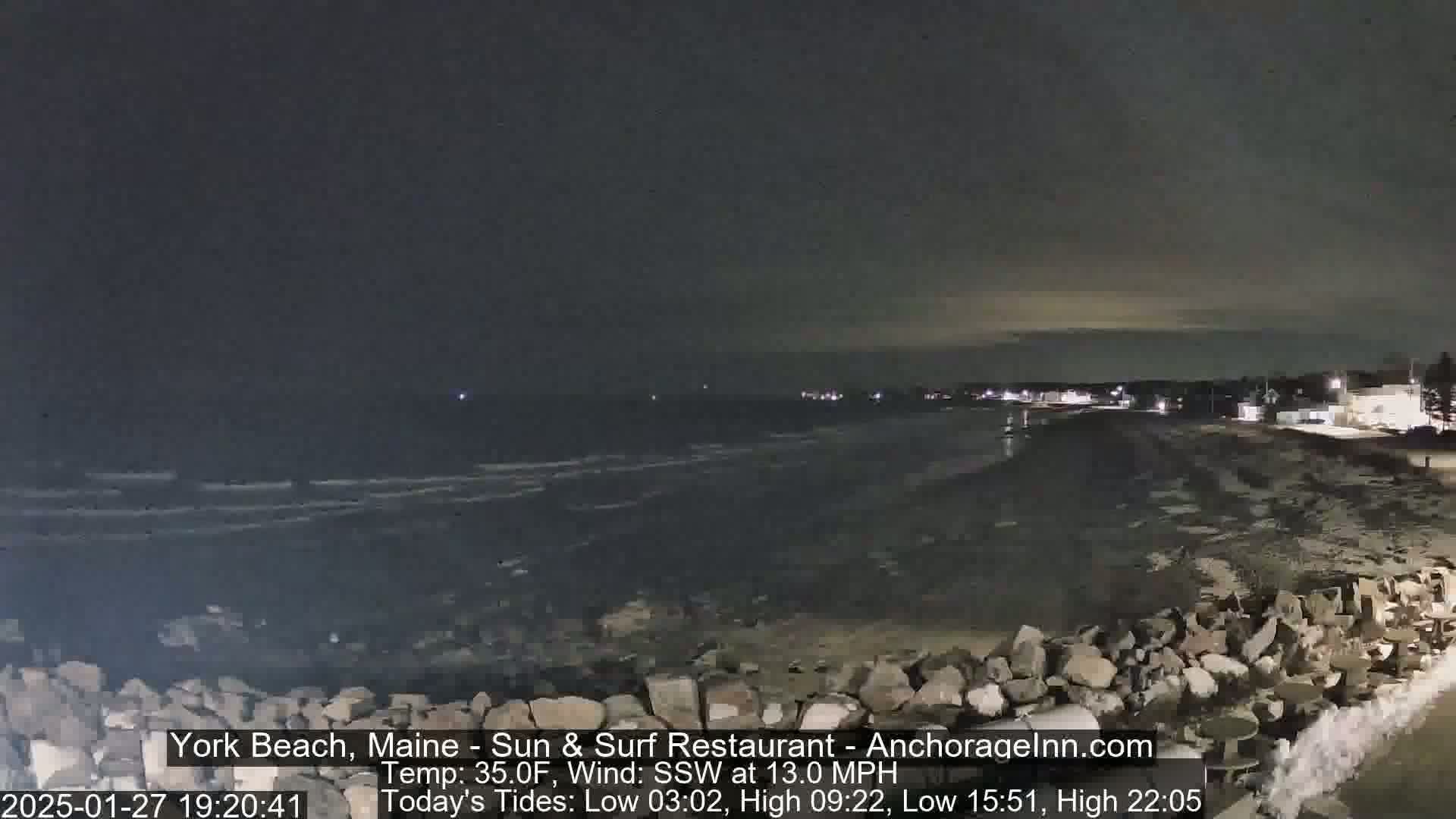 A dark nighttime beach scene shows a rocky shoreline with dark, choppy water under a mostly clear sky, with a gentle wind blowing from the southwest at 13 miles per hour.