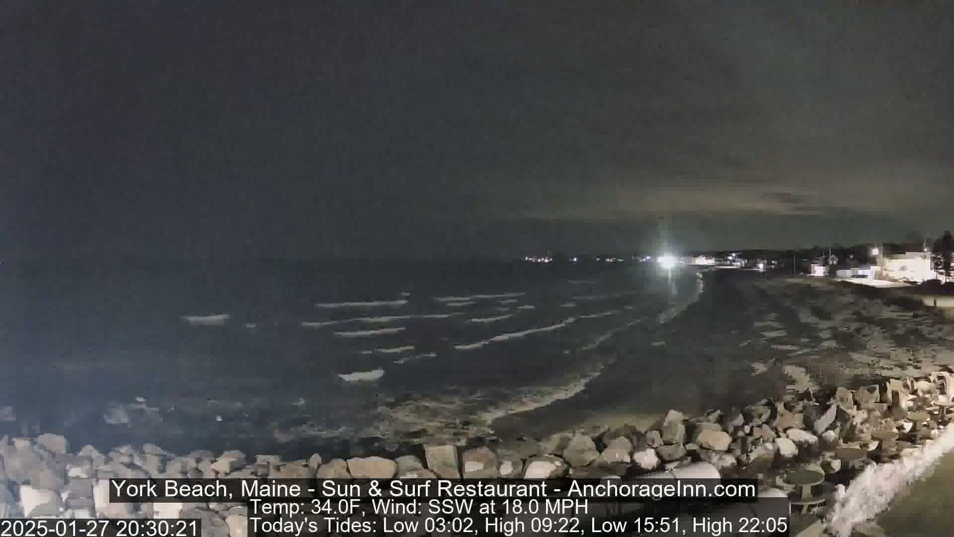 A nighttime coastal scene shows dark, wavy water lapping against a rocky shore under a cloudy sky with an 18 mph wind.