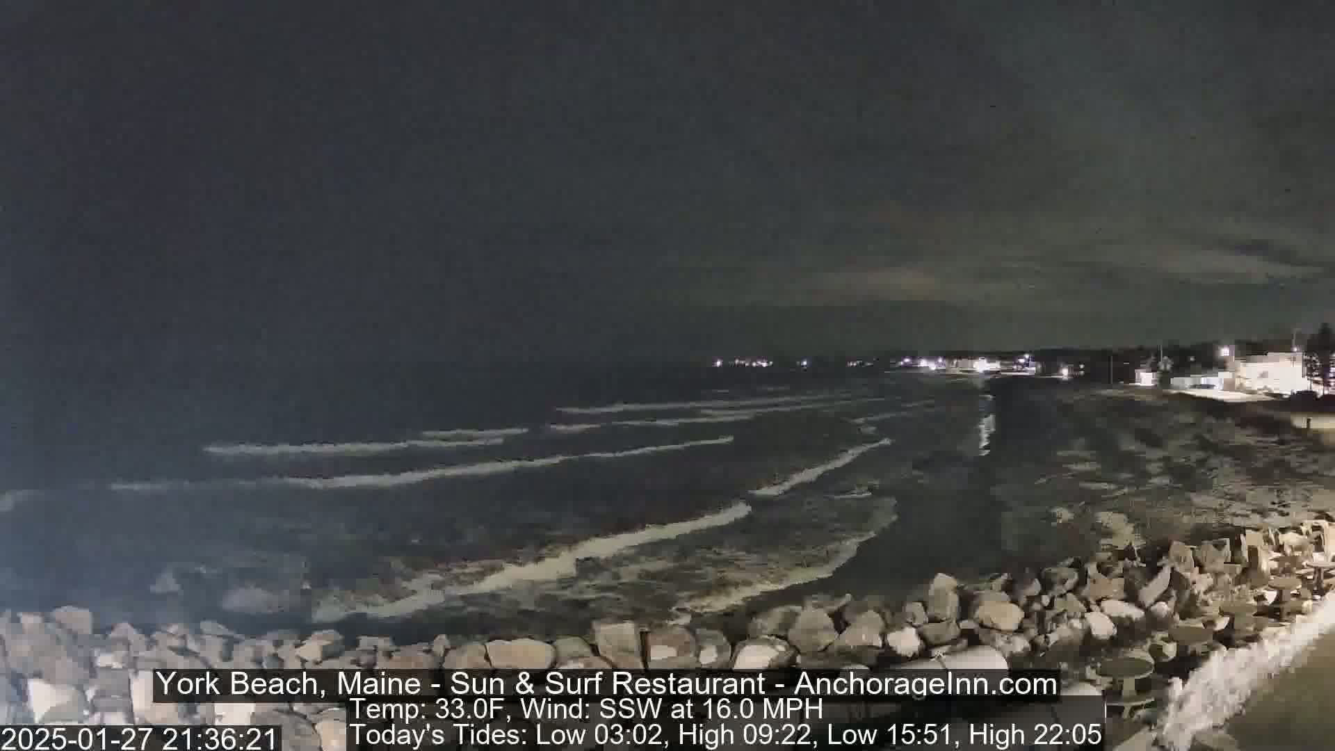 A nighttime coastal scene shows dark, wavy water lapping against a rocky shoreline under a mostly clear sky, with a 16 mph south-southwesterly wind and a temperature of 33°F.