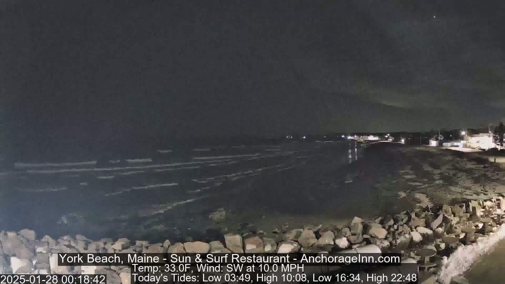 A nighttime coastal scene shows a dark, wavy ocean meeting a rocky shore under a clear, dark sky.