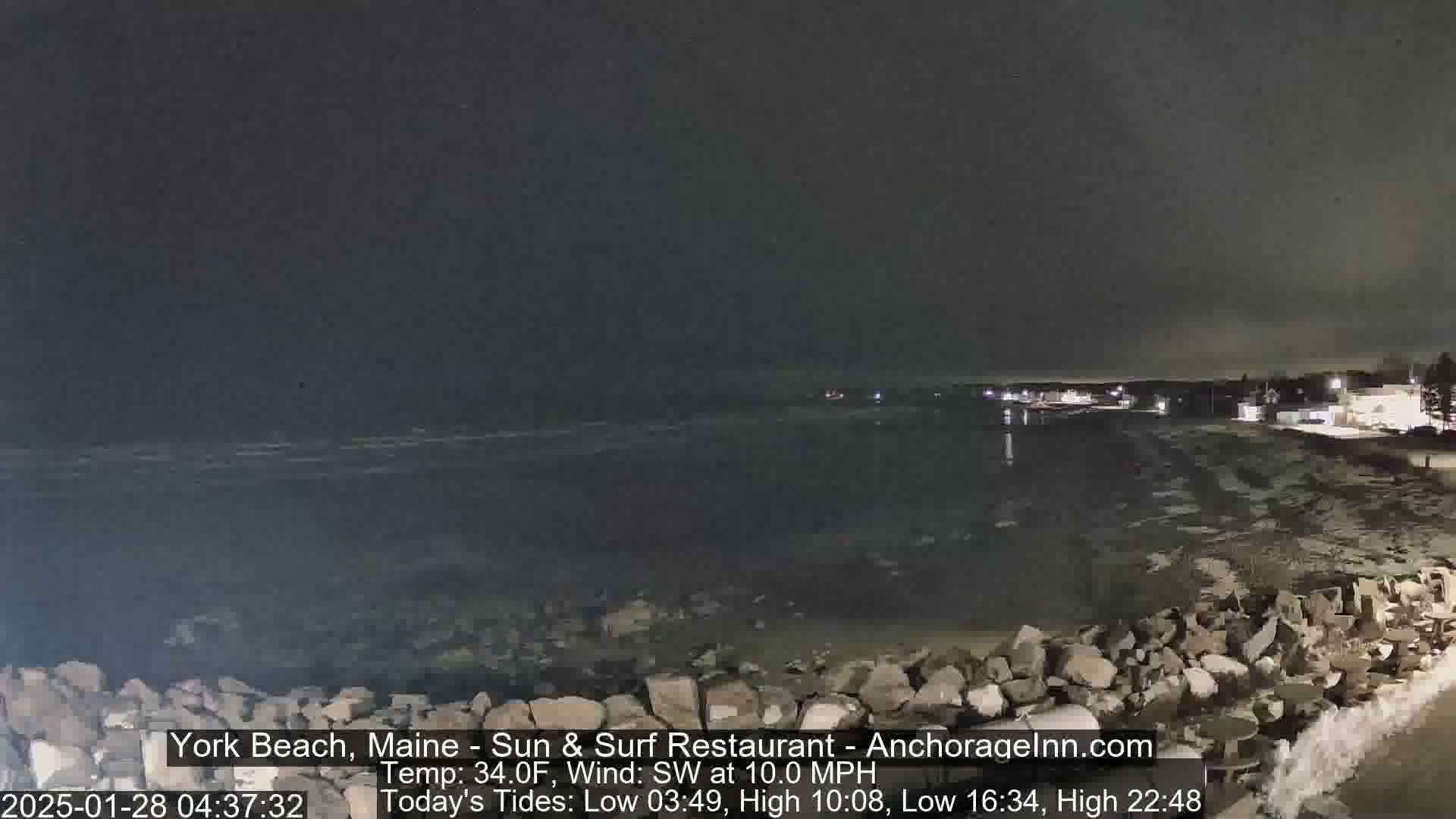 A dark, nighttime view of a rocky shoreline and calm ocean under a cloudy sky.