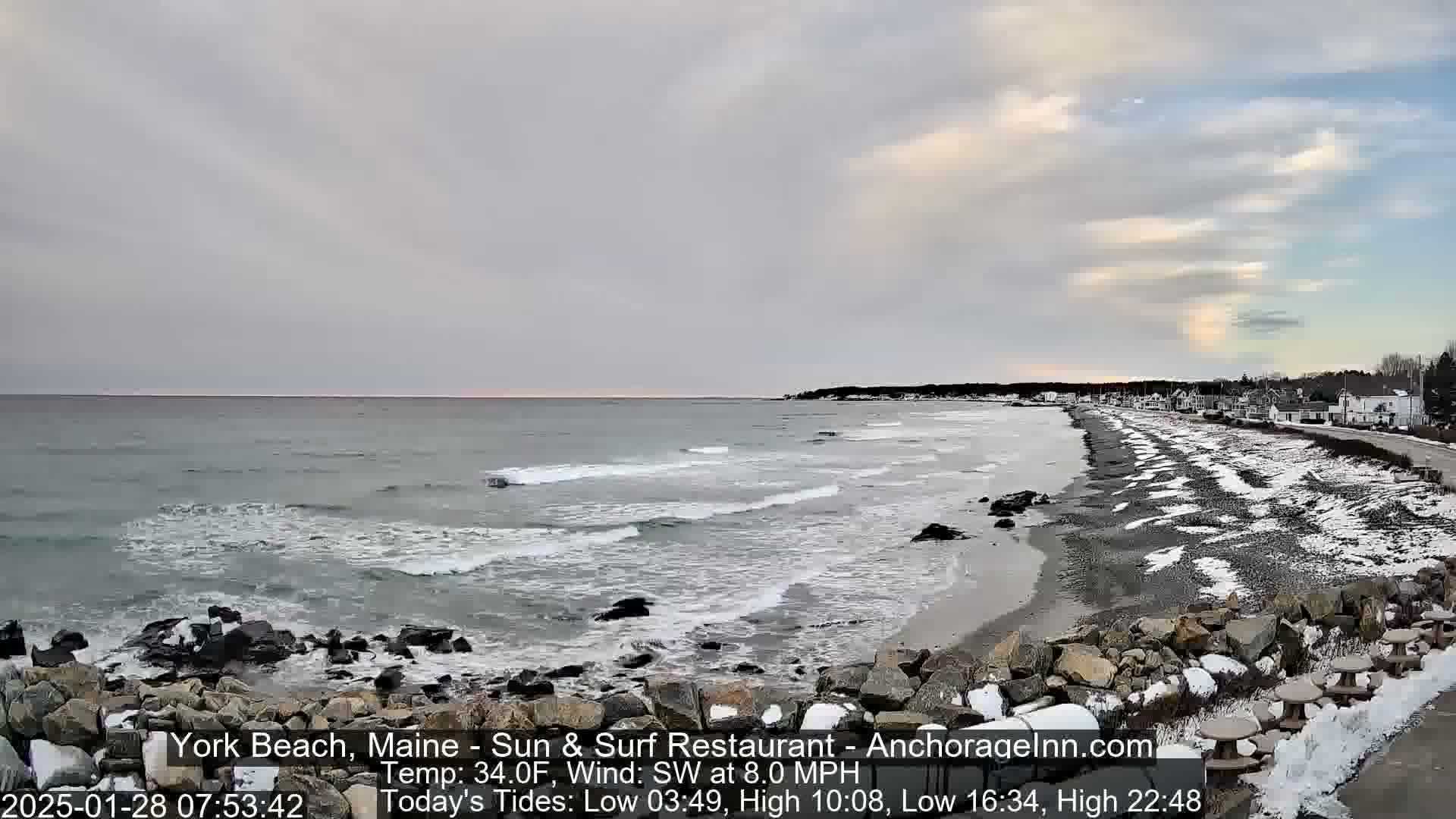A partly cloudy sky hangs over a snow-dusted beach with a rocky shoreline and gentle waves lapping the shore.