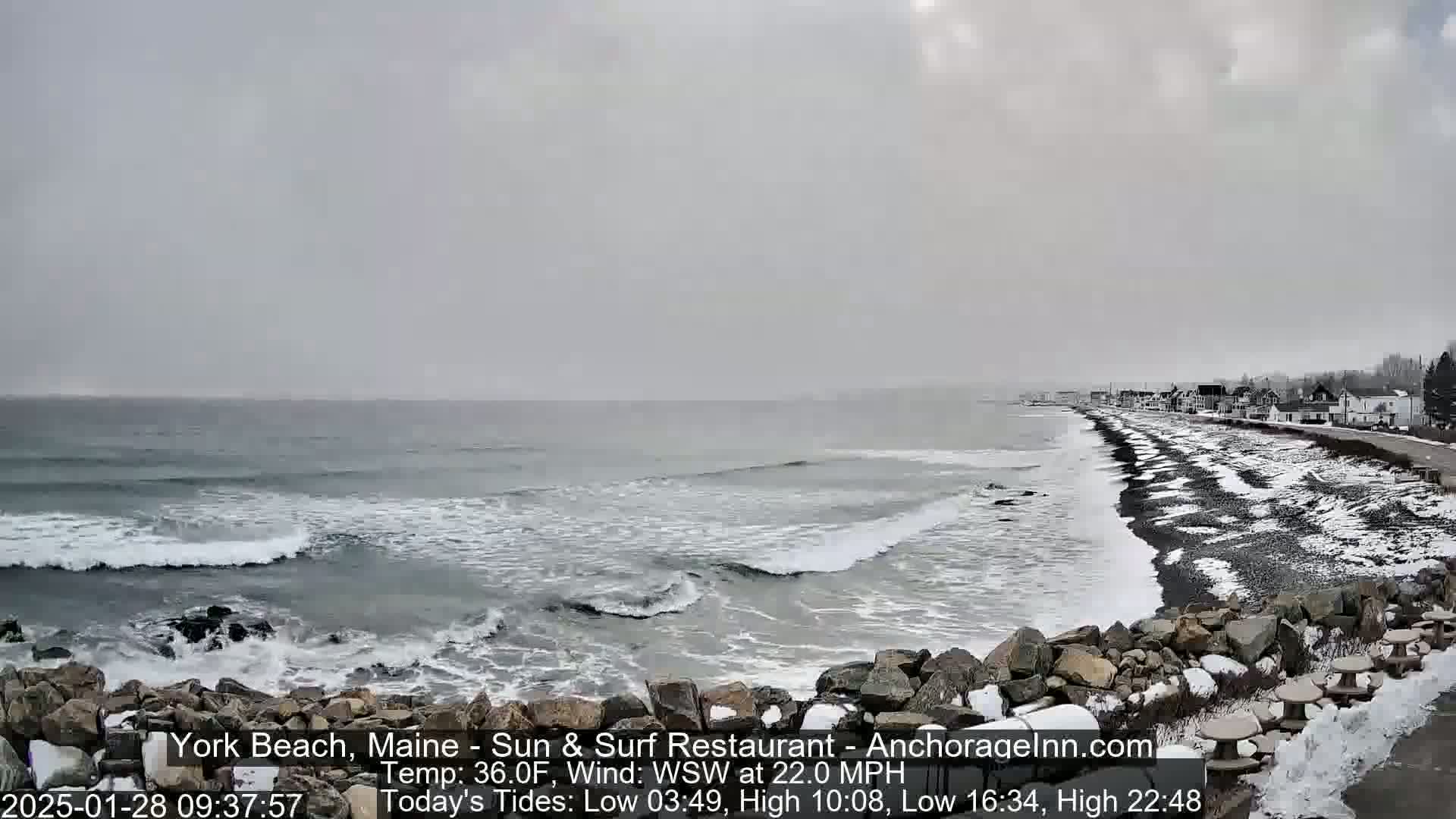 A snow-covered beach with a rocky shoreline and houses in the distance is shown under an overcast sky, with waves breaking on the shore.