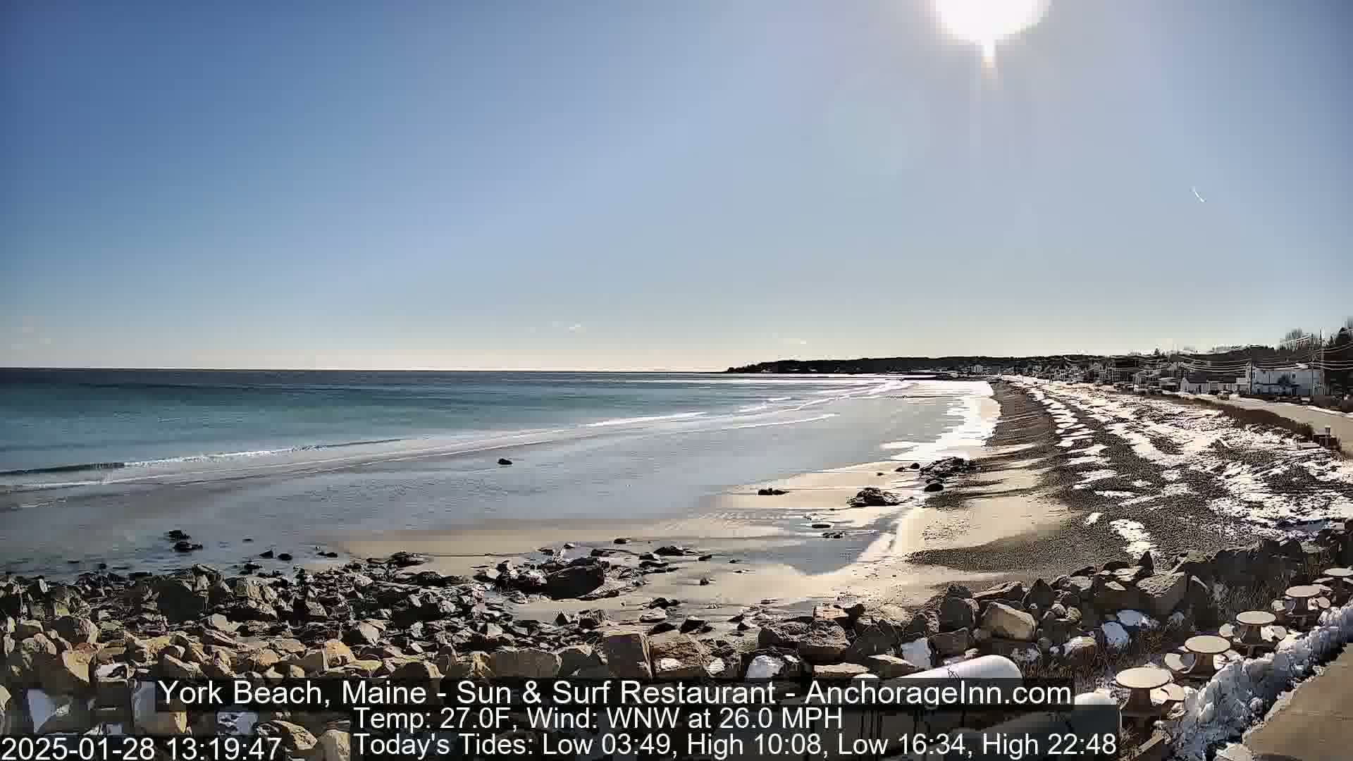 A sandy beach with some snow patches stretches along a rocky shoreline under a bright, sunny sky, with a calm ocean in the background.