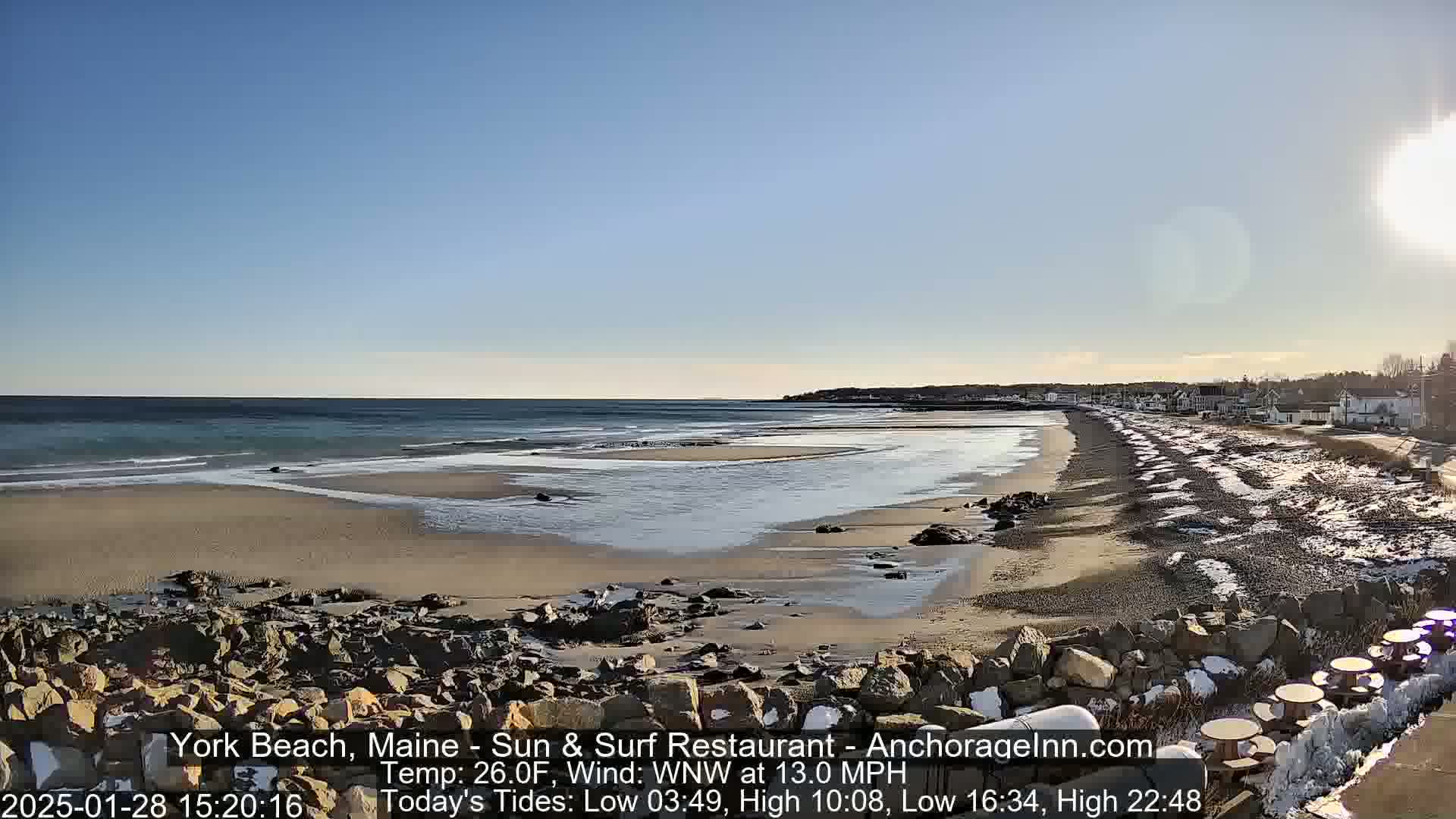 A sunny, cold day at a beach with a partly frozen tidal area, rocky shoreline, and buildings in the distance.