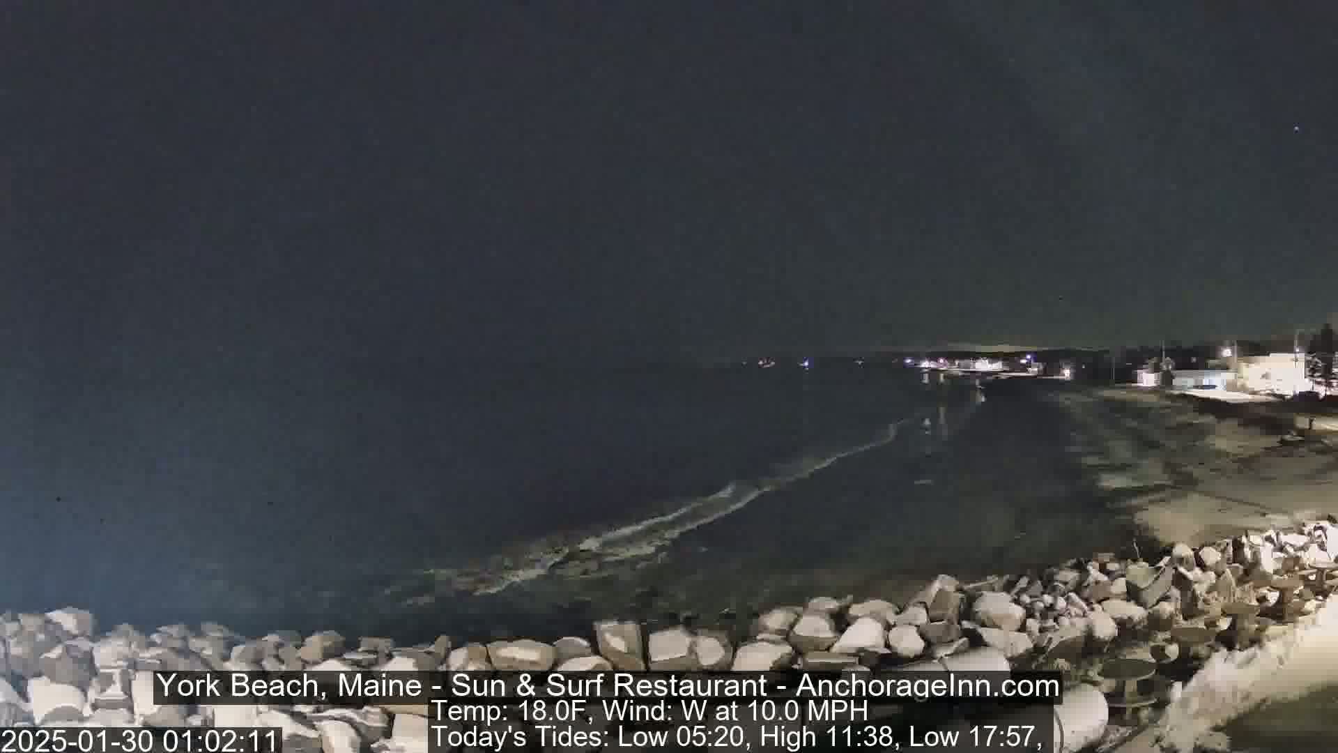 A dark, nighttime view of a rocky shoreline and ocean, with a slight wind and calm waves.