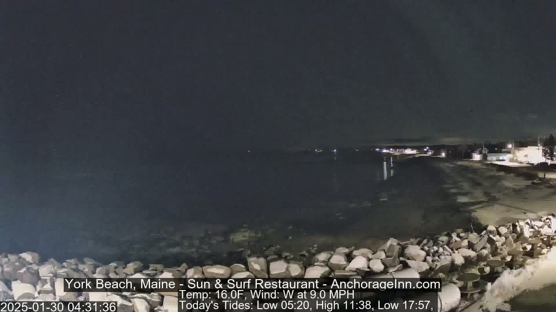 A nighttime coastal scene shows a dark body of water, a sandy shore, and a rocky barrier, under a clear, dark sky.