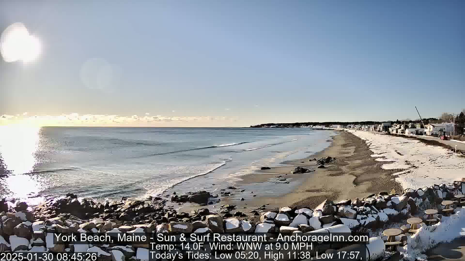 A partially snowy beach with calm waves and a clear, sunny sky stretches out to a distant coastline.