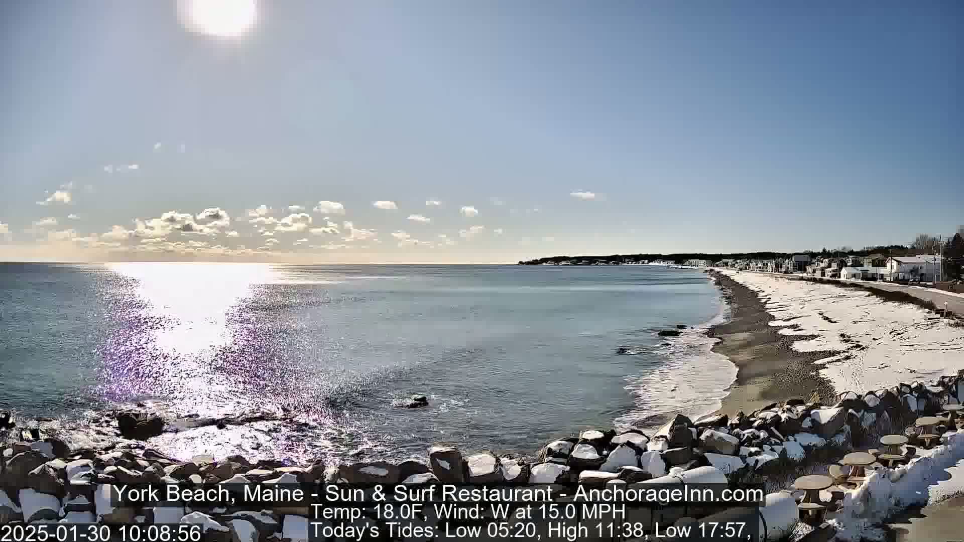 A partly sunny, cold day shows a partly snow-covered beach with a calm ocean, and a shoreline with buildings visible in the distance.