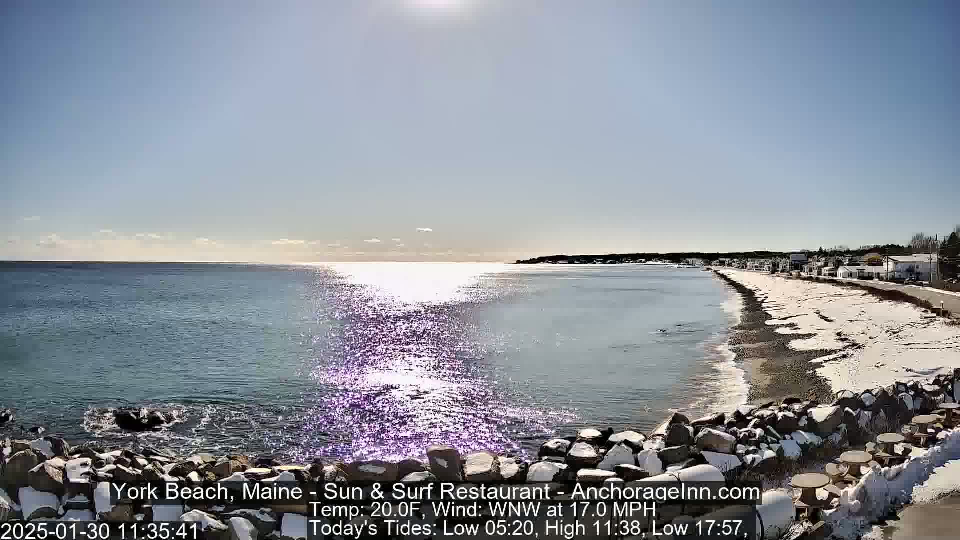 A partly sunny, cold day at a snow-dusted beach shows a calm sea reflecting sunlight, with a shoreline lined with buildings and rocky barriers.