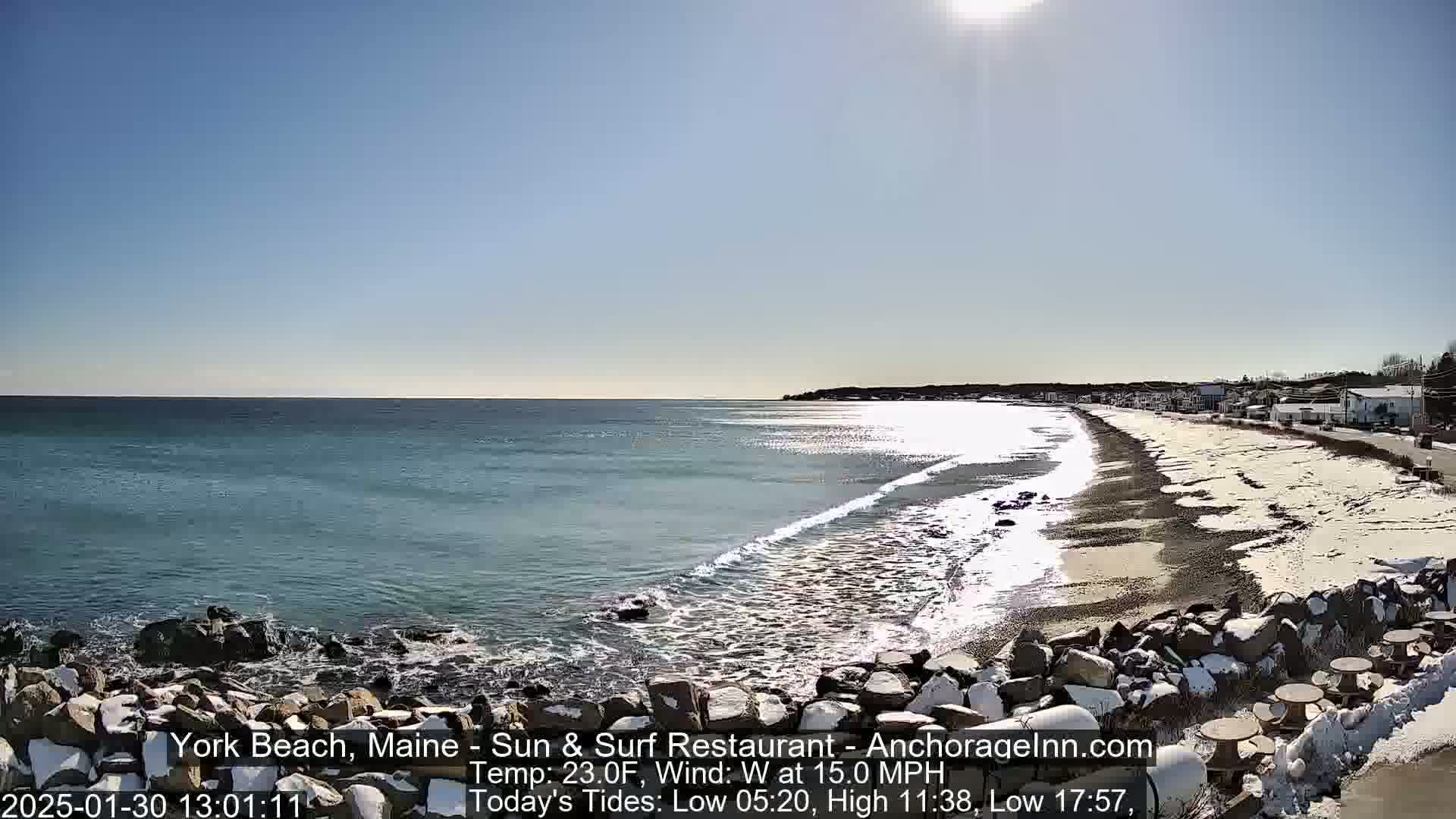 A sunny, cold day at a beach with a rocky shore, partially covered in snow, shows gentle waves lapping the sandy beach where some buildings are visible in the distance.