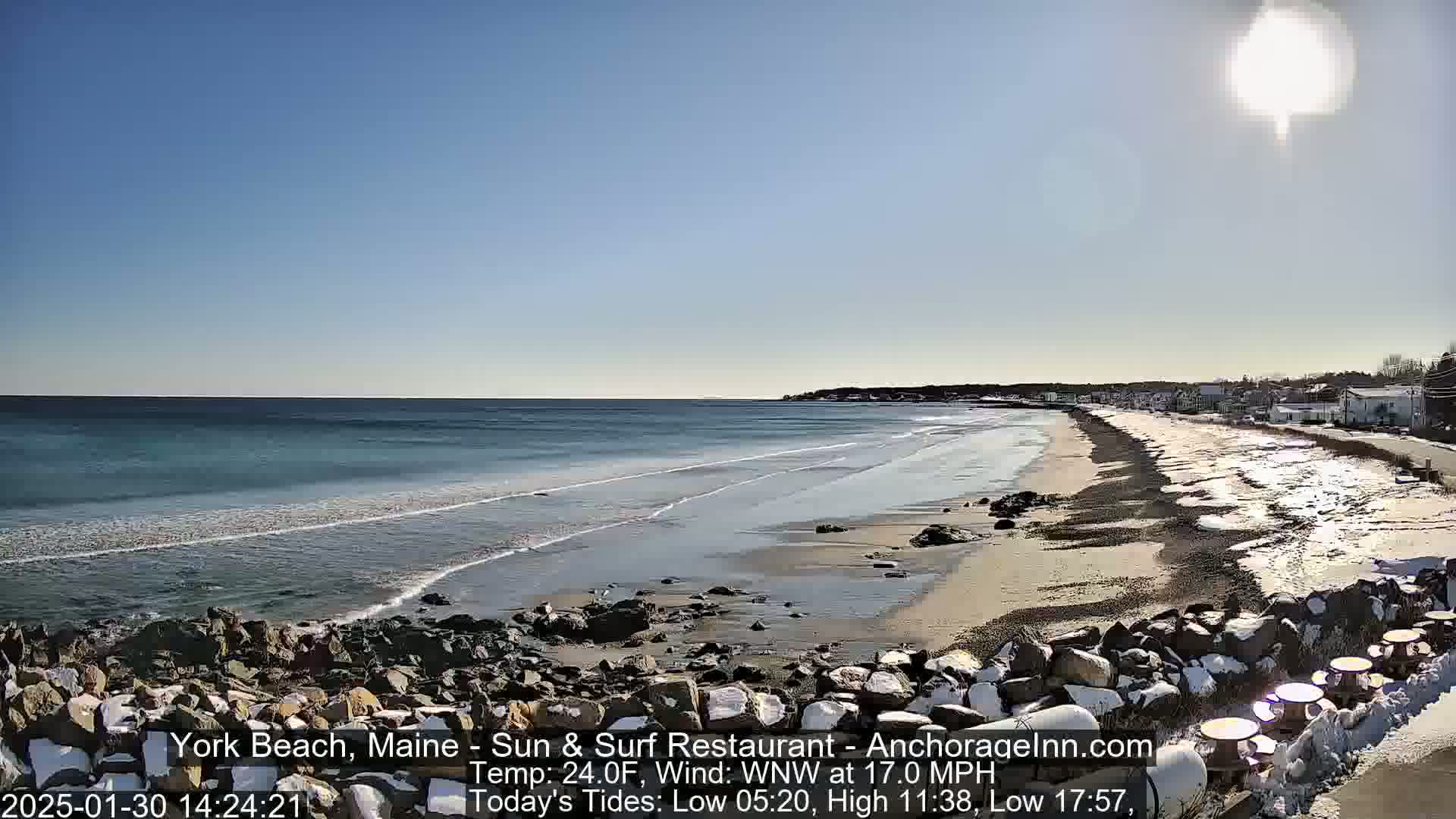 A sandy beach with a rocky shoreline and calm ocean waves stretches under a bright, sunny sky; snow patches are visible on the rocks and the upper beach.