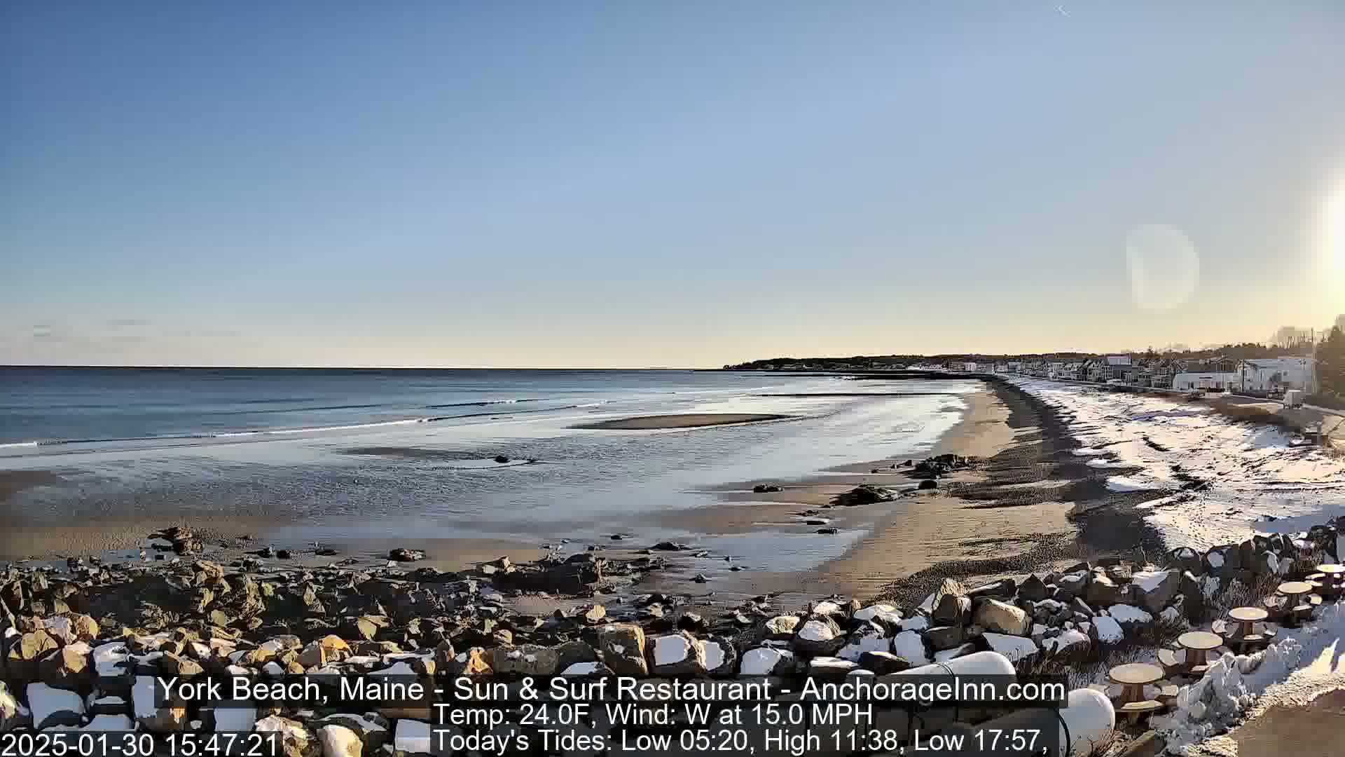 A sandy beach with some snow and ice, partially rocky shoreline, and a calm ocean under a clear, sunny sky.