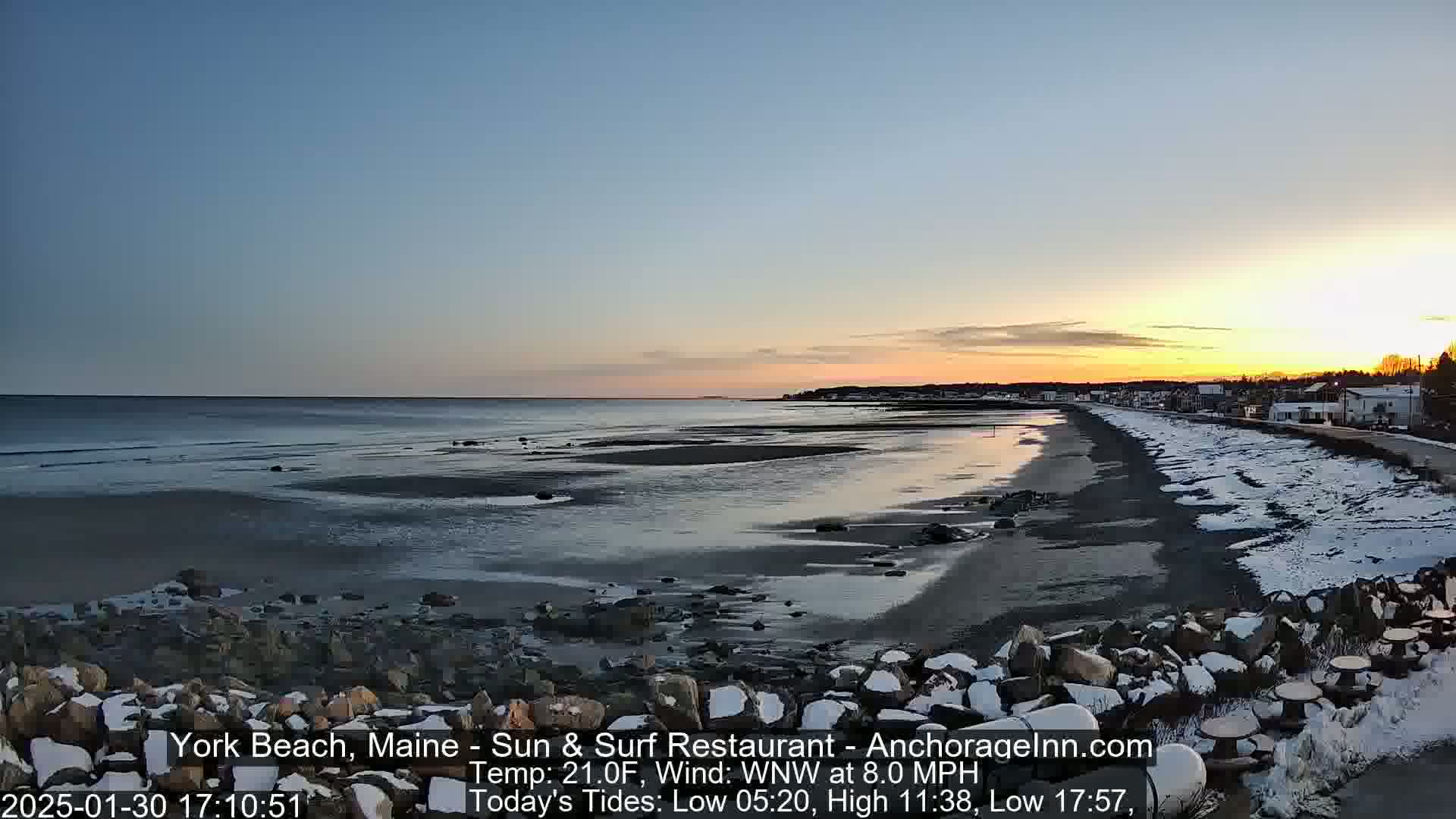 A partially snow-covered beach at sunset, with a calm ocean and a town visible in the distance.