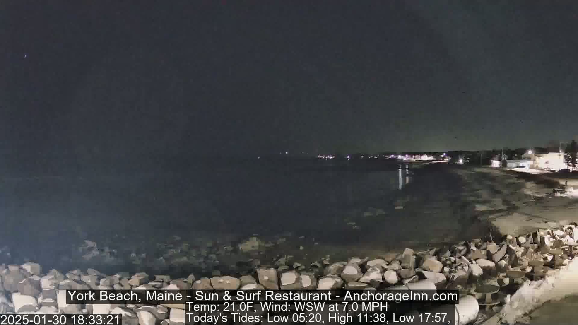 A nighttime view of a rocky shoreline and calm water, with distant lights visible across the water under a clear, dark sky.