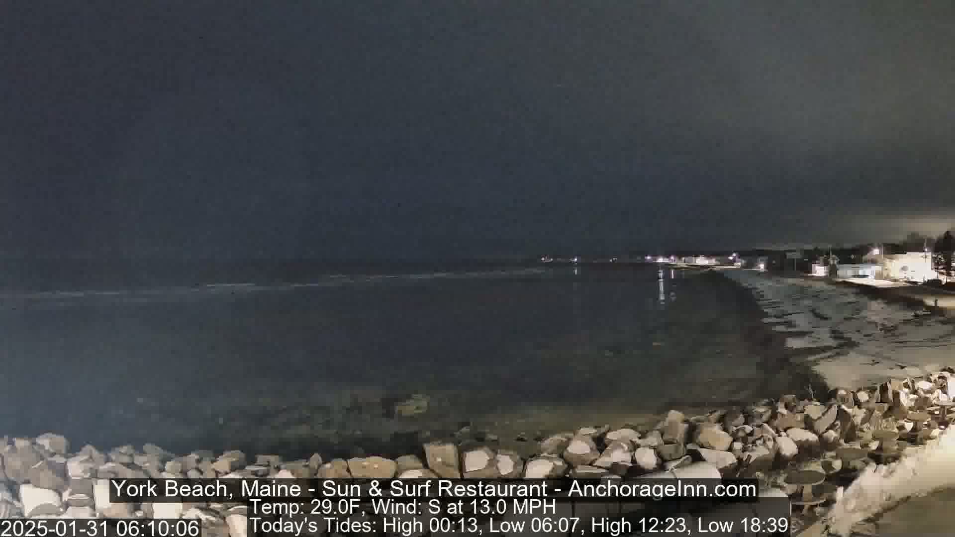 A nighttime view of a dark, rocky beach and calm ocean under a cloudy sky.