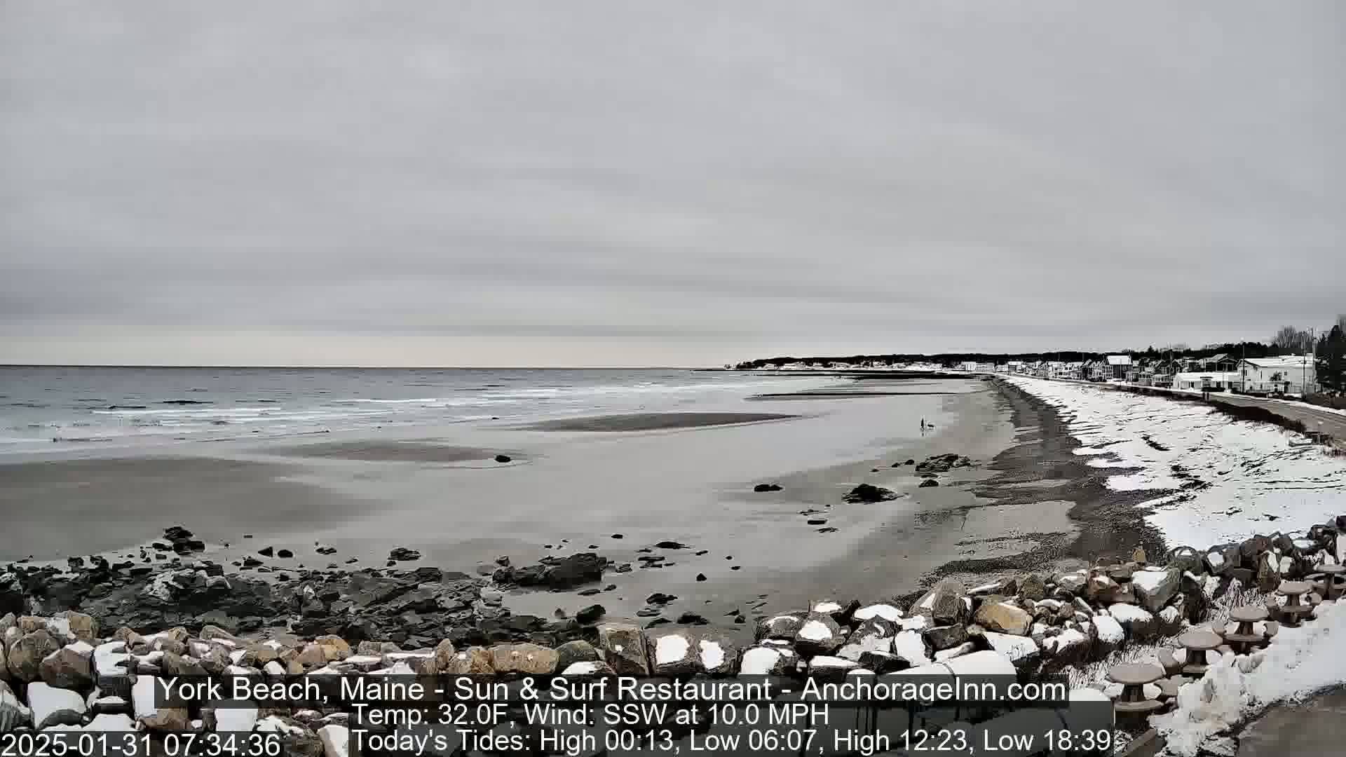 A snow-dusted beach stretches along a gray, overcast sky, with a calm sea and a few buildings visible in the distance.