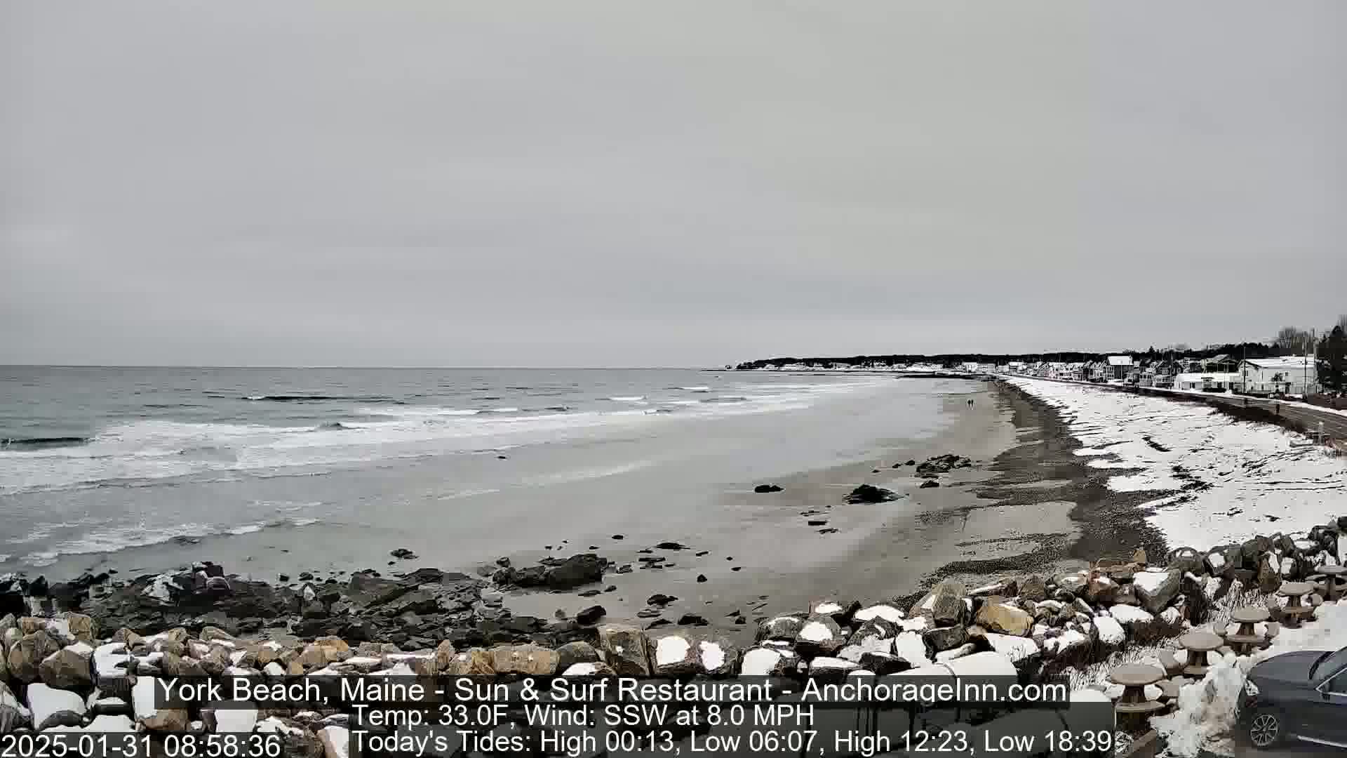 A snow-dusted beach stretches along a gray, overcast ocean, with a few people walking along the shore and buildings visible in the distance.