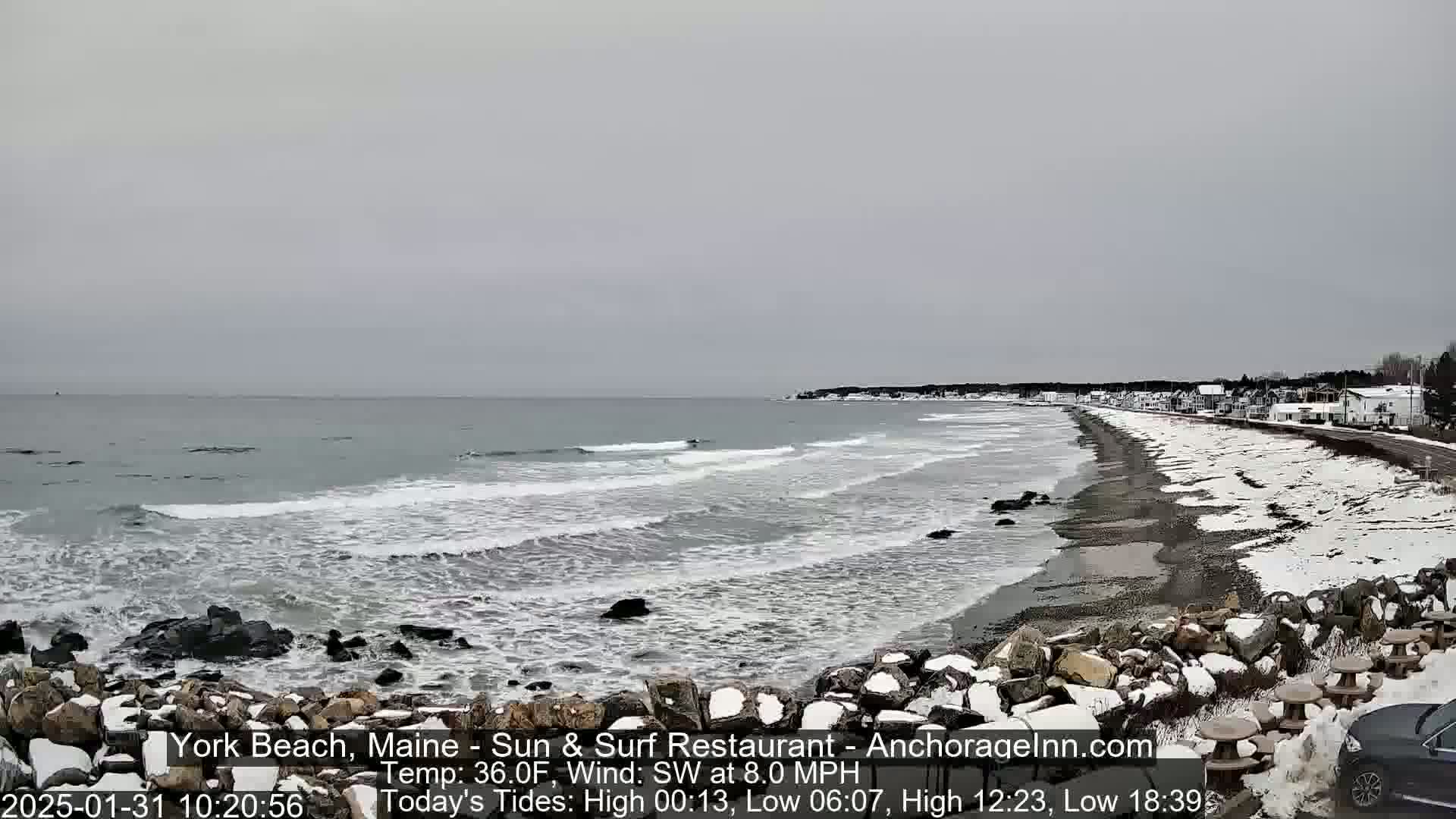 A snow-dusted beach with gray, overcast skies and gentle waves rolls onto a shoreline with a few buildings in the distance.