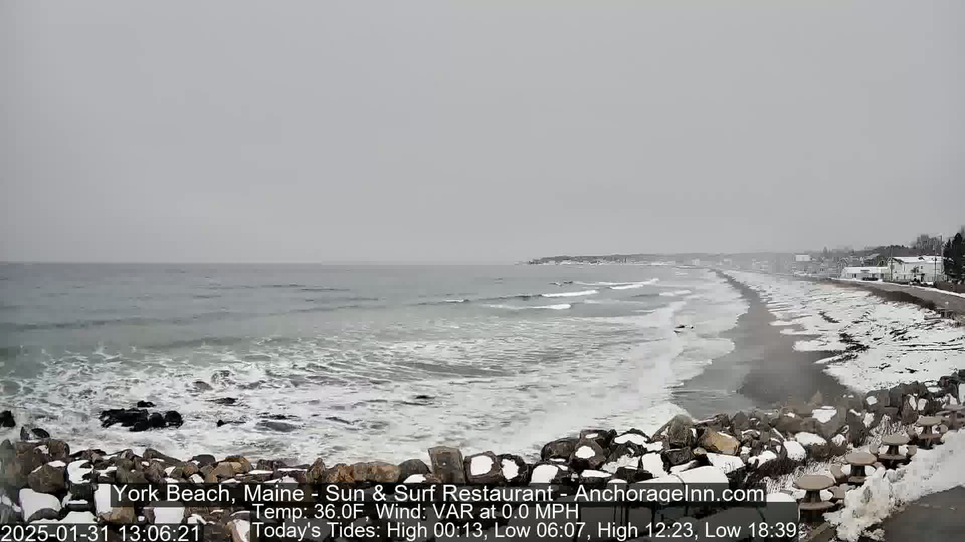 A snow-covered beach with a calm ocean and distant buildings under an overcast sky.