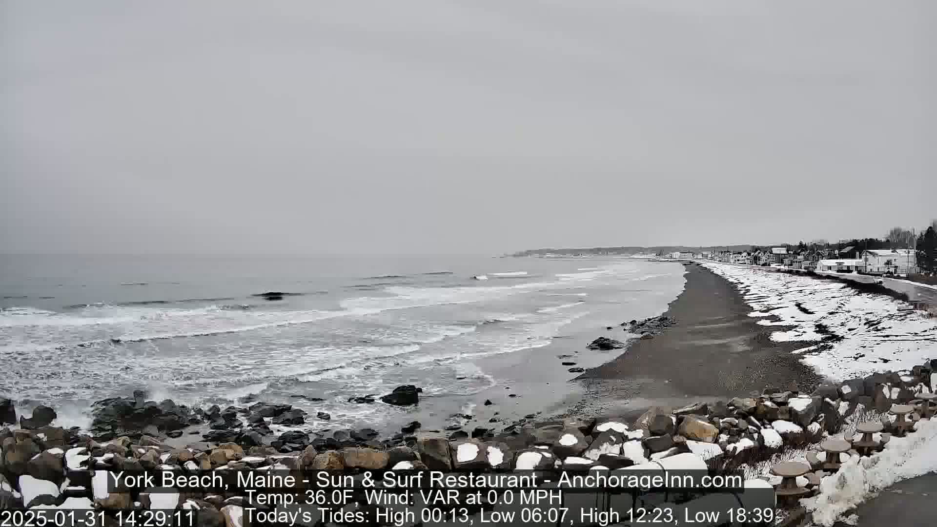 A snow-dusted beach stretches alongside a calm, grey ocean under an overcast sky.