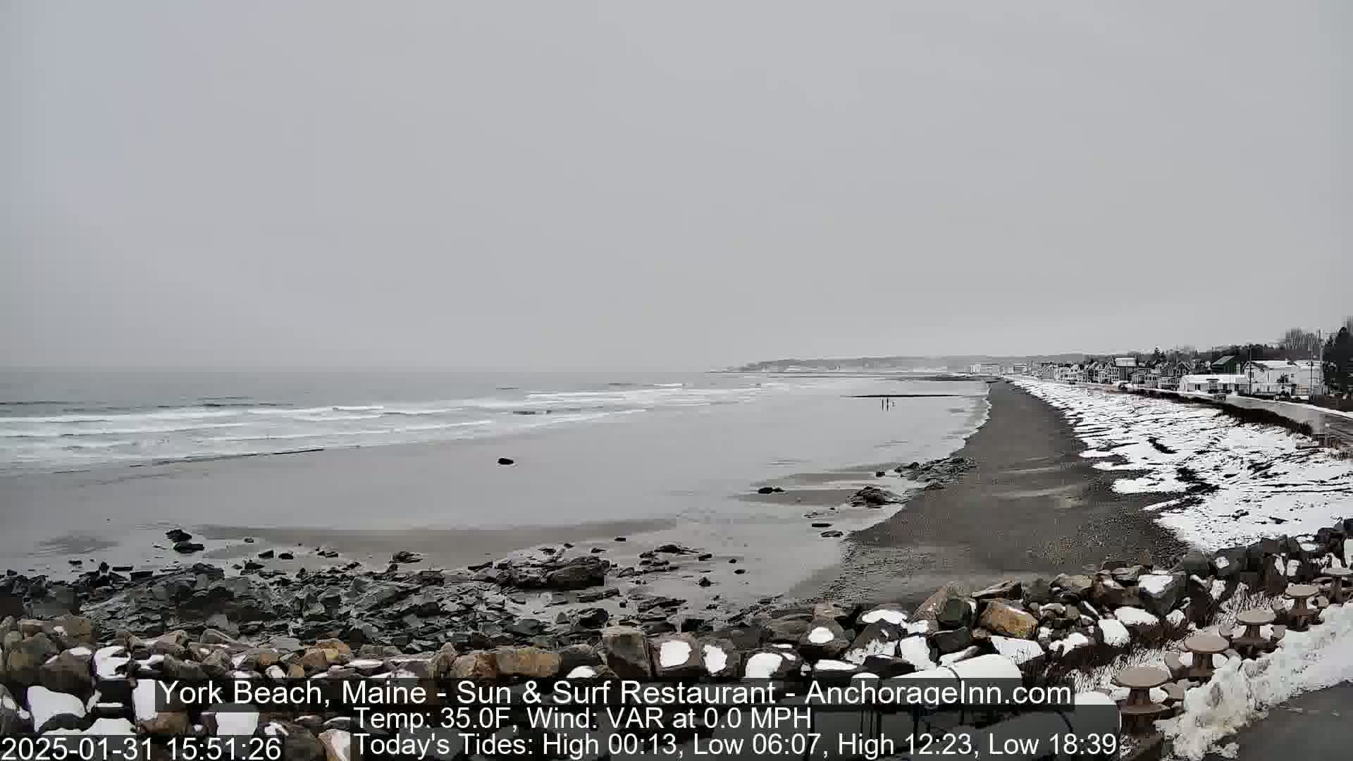 A snow-dusted beach stretches along a calm, gray ocean under an overcast sky.