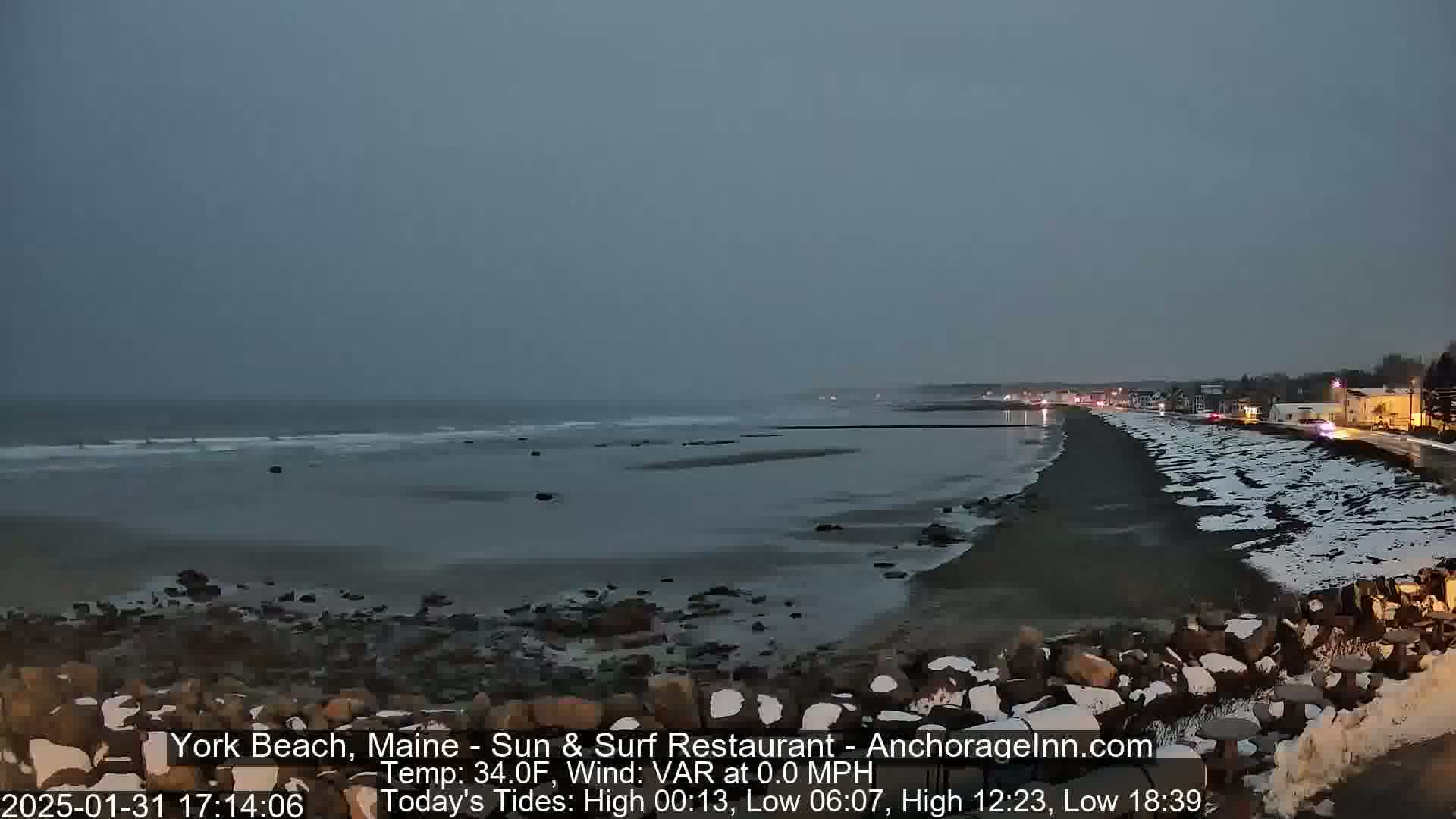 A snow-dusted beach at twilight, with a calm ocean and a town visible in the distance under a gray sky.