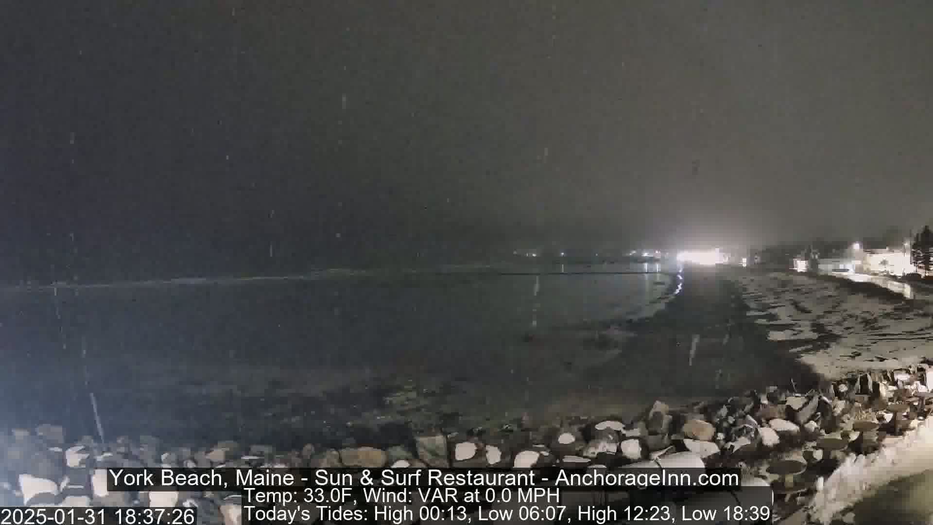 A nighttime snow flurry falls over a dark, rocky shoreline and calm body of water, with distant lights visible.