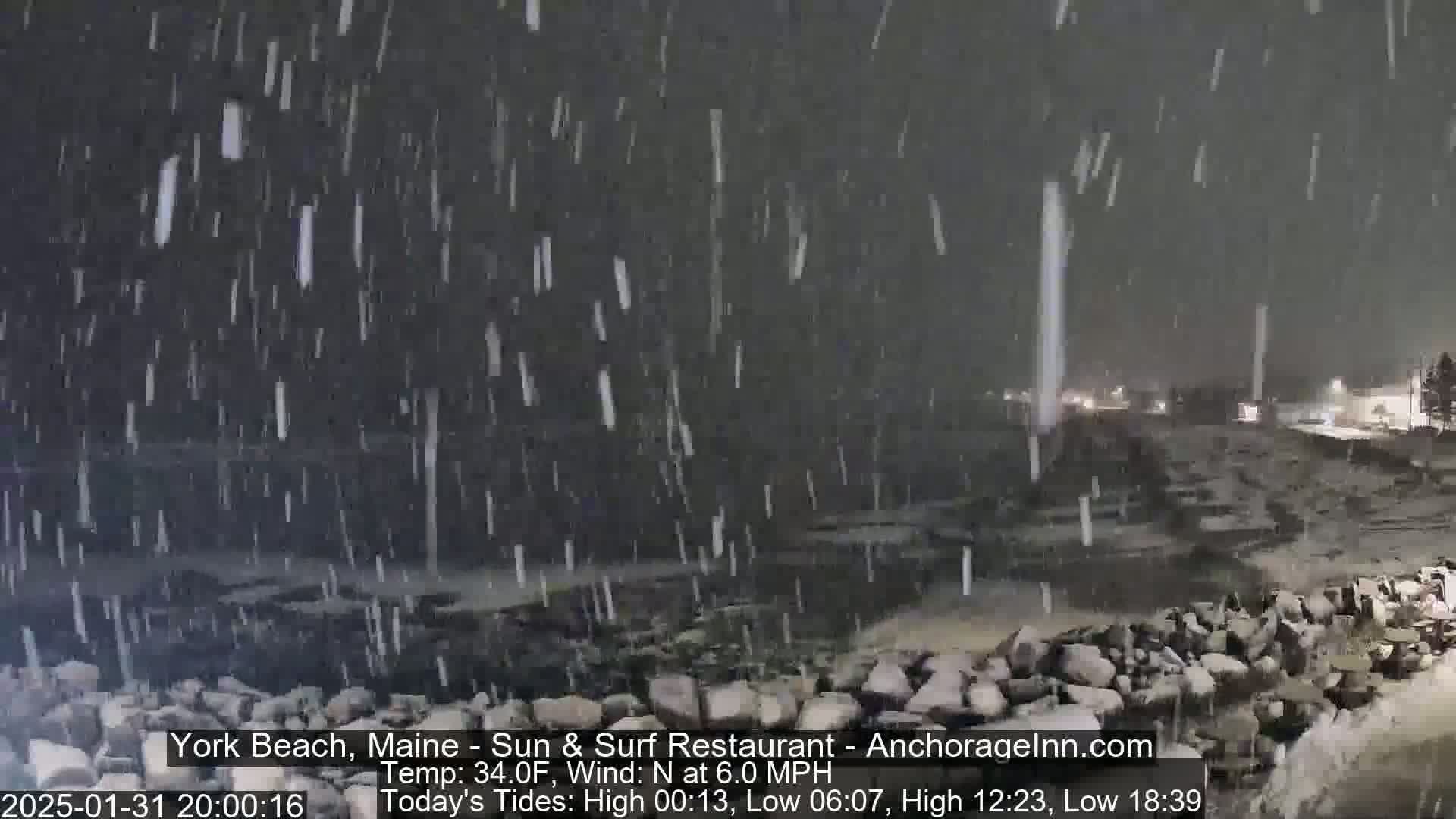 A nighttime scene of snow falling heavily over a rocky shoreline, with lights visible in the distance.