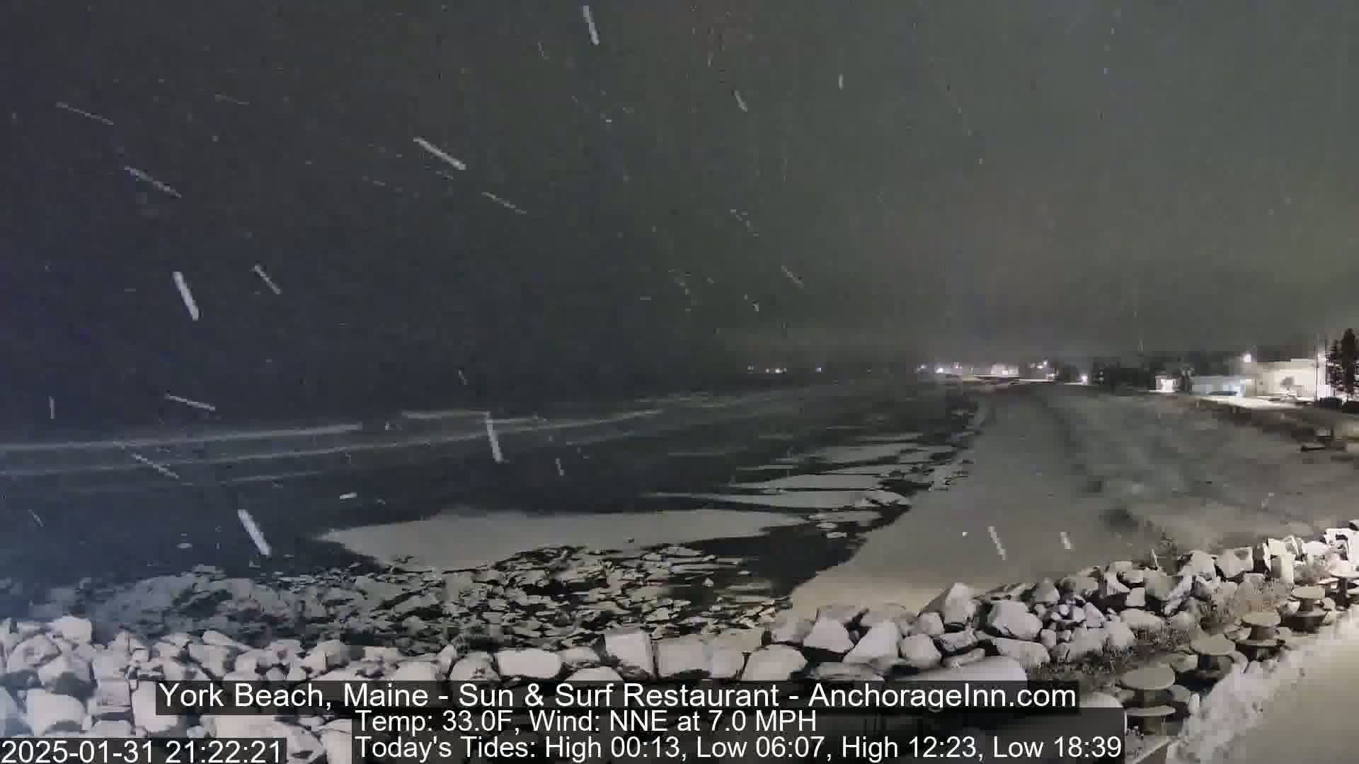 A snow-covered beach at night with a dark body of water partially frozen over, under a falling snow.