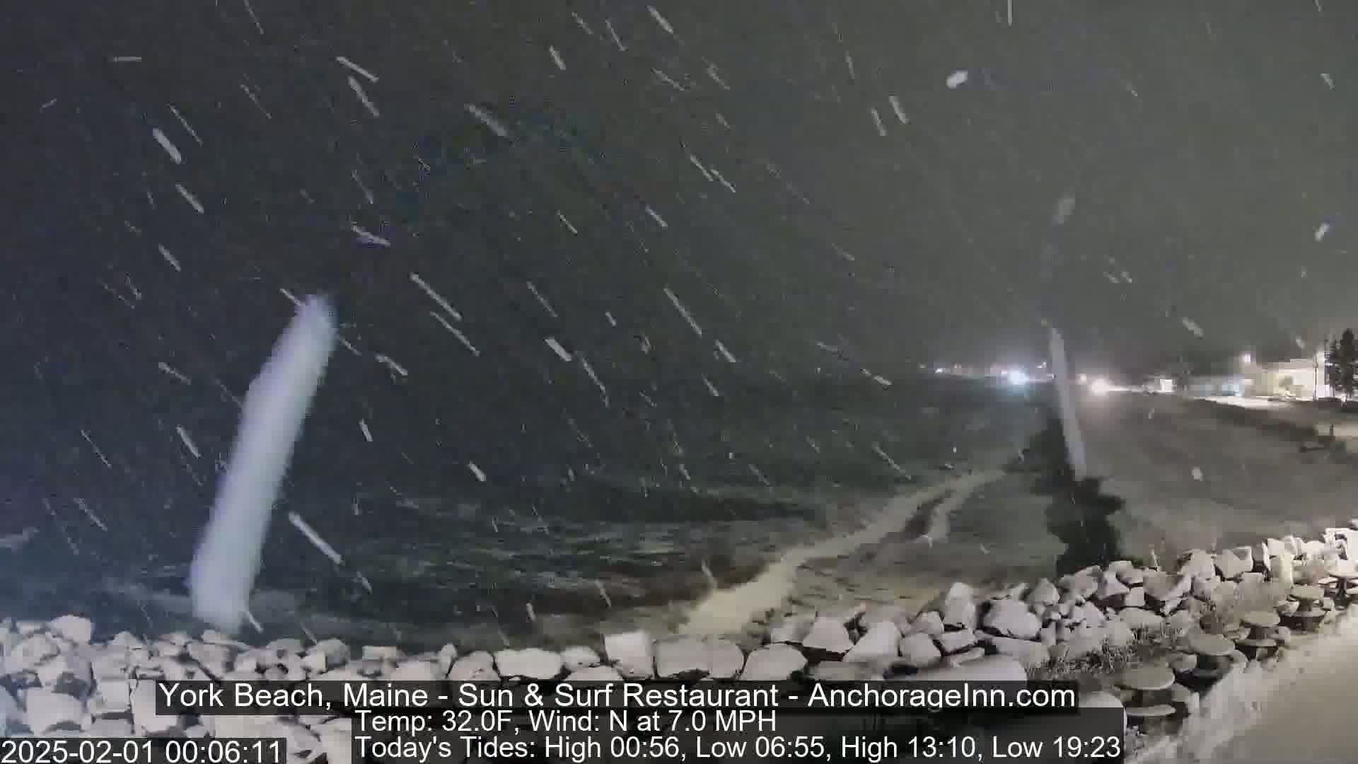 A nighttime scene of a snow-covered rocky shoreline with waves crashing against it during a heavy snowfall.