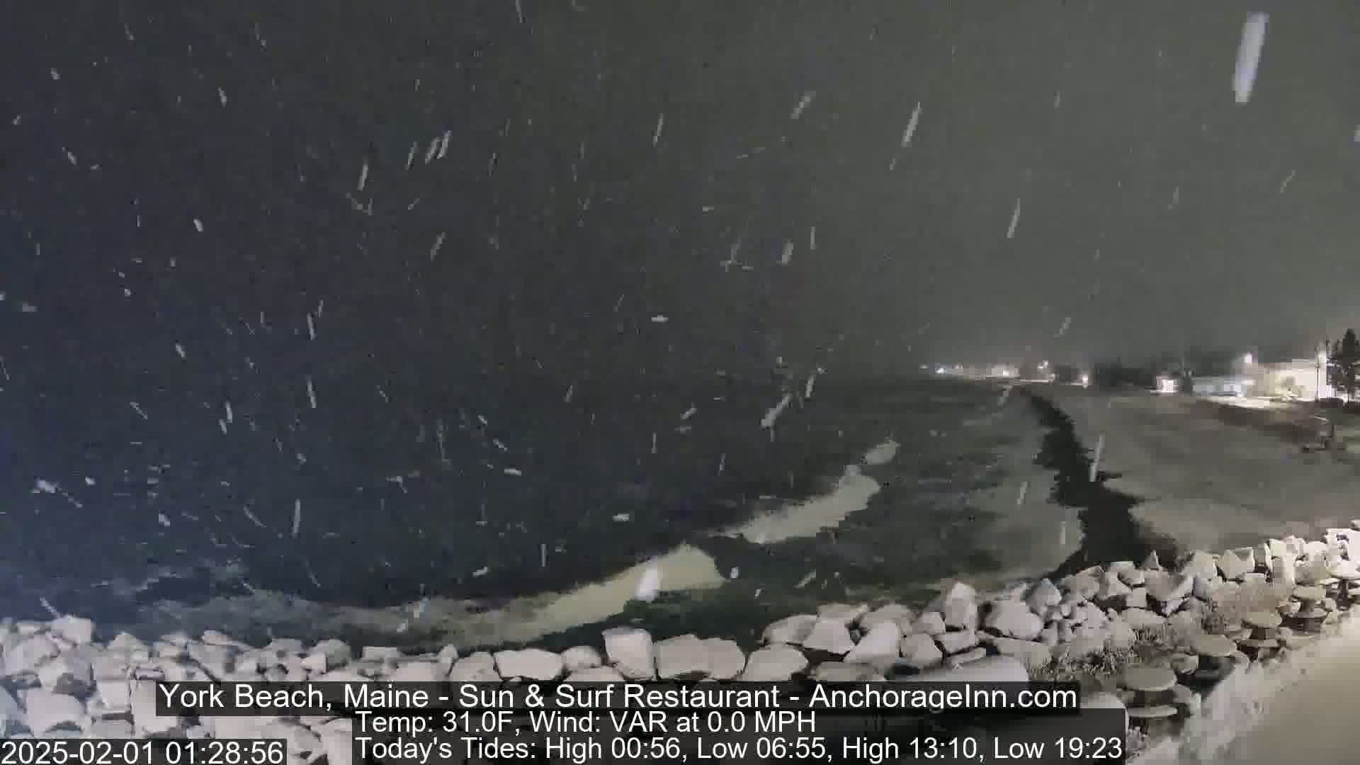 A nighttime snowstorm falls over a rocky shoreline and dark, icy water.