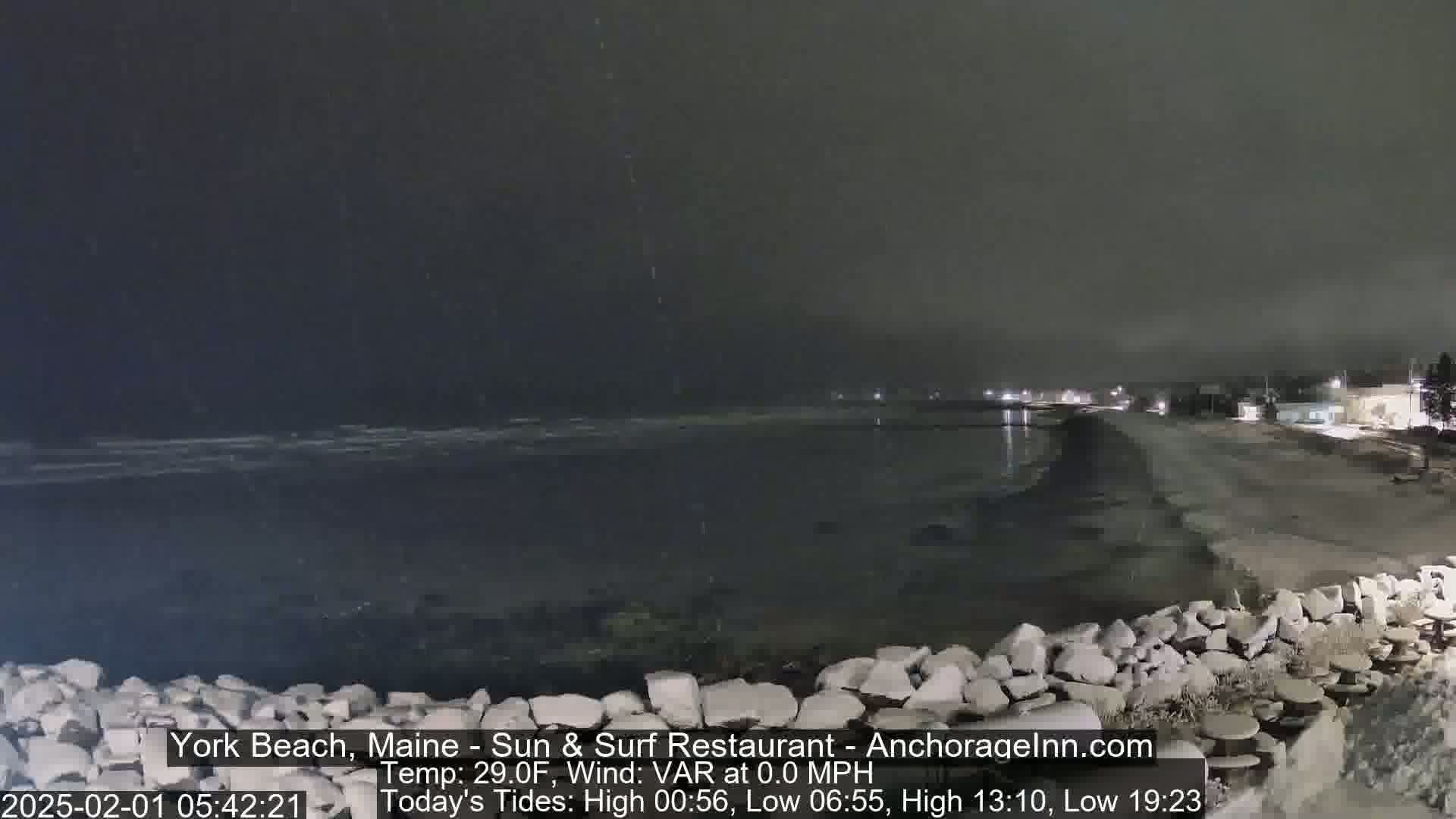 A dark, nighttime beach scene shows a calm ocean with a rocky barrier in the foreground under snowy conditions.