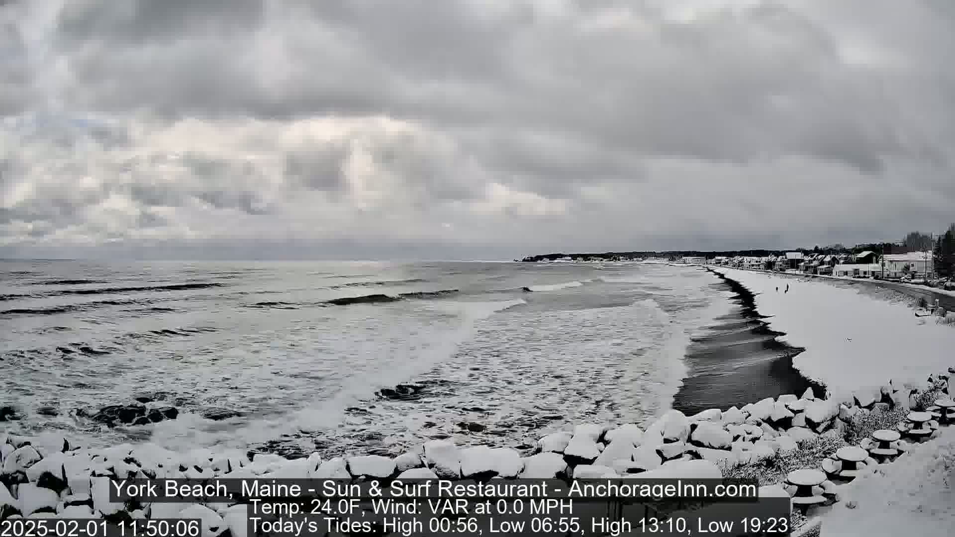 A snow-covered beach with dark, wavy ocean and overcast skies is seen from a high vantage point.