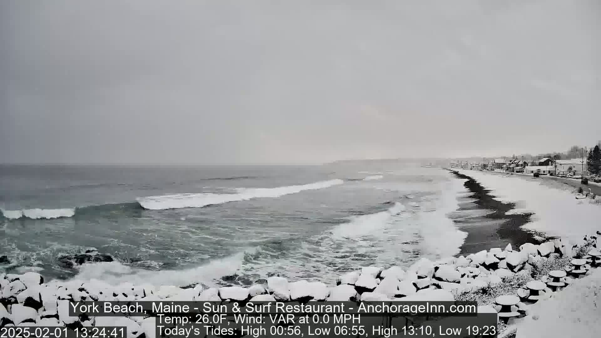 A snow-covered beach with dark sand and a rocky shore is viewed from a slight elevation showing a grey, overcast sky and waves rolling onto the beach.