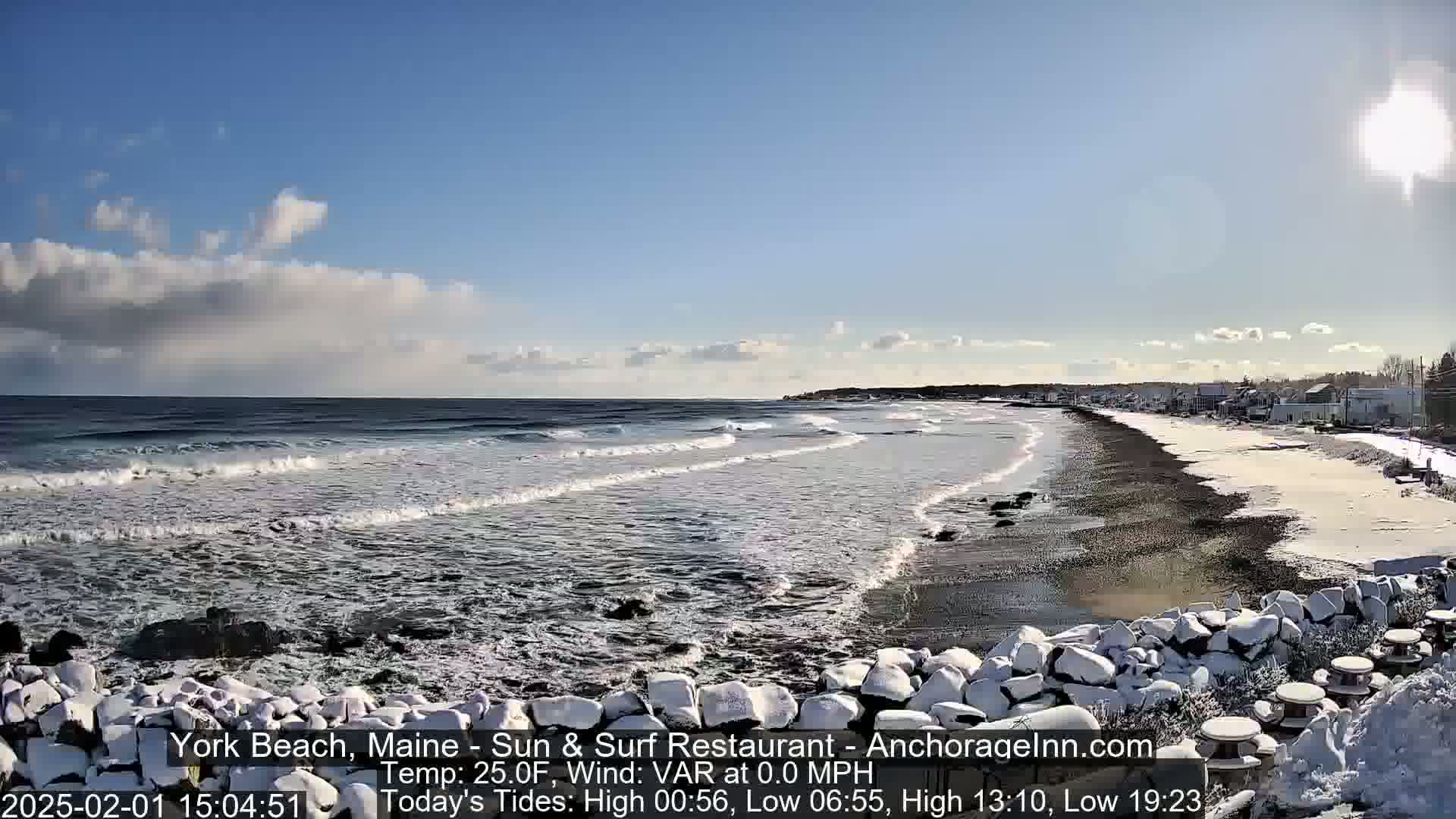 A partially sunny day shows a snow-dusted beach with gentle waves lapping the shore, some buildings visible in the distance.