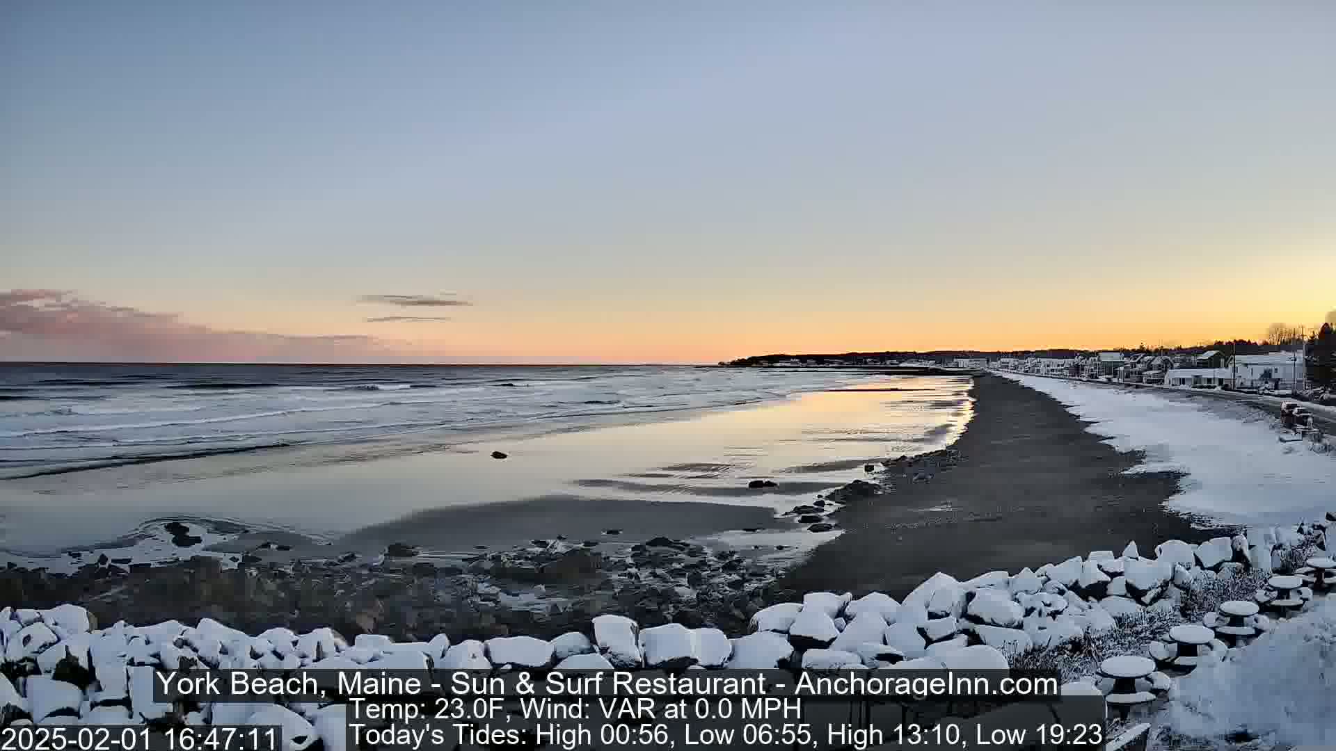 A snow-dusted beach at sunset shows calm waves lapping the shore, with a town visible in the distance.