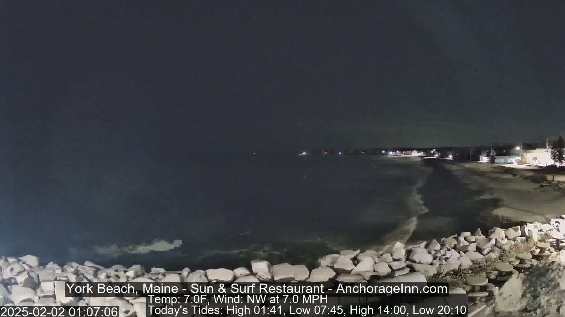 A dark, nighttime beach scene shows gentle waves lapping against a rocky shoreline under a clear, dark sky.