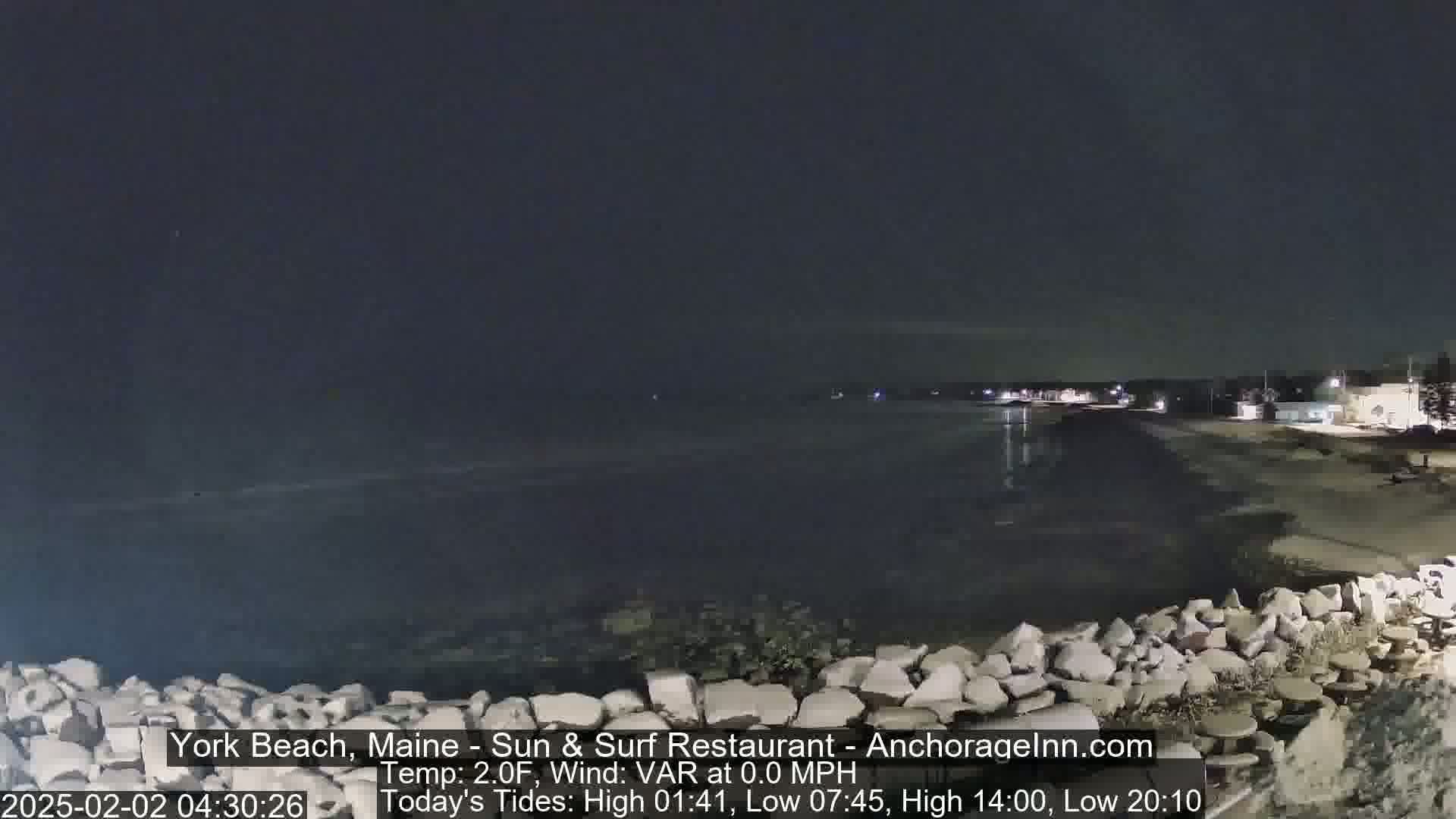A dark nighttime view of a calm ocean shore with a rocky barrier in the foreground, under a clear sky.