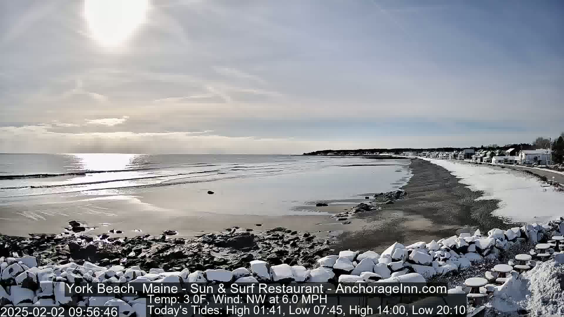 A snow-covered beach with calm waves and a partly sunny sky stretches toward a distant shoreline with buildings.