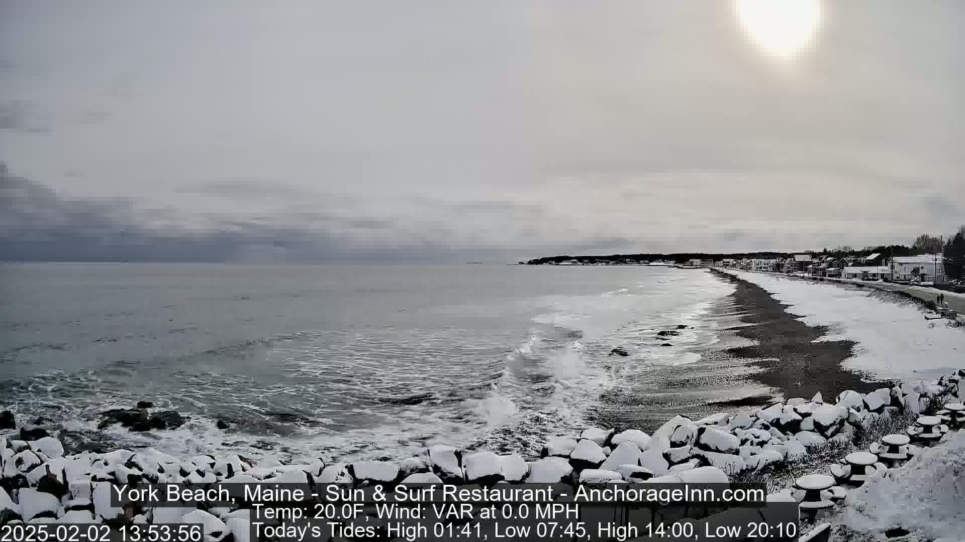 A snow-covered beach stretches along a dark, cold ocean under a mostly cloudy sky with a bright sun.