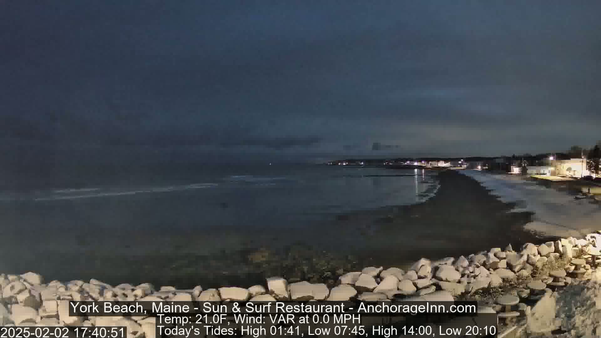 A dark, nighttime beach scene shows a calm, dark ocean under a cloudy sky, with a rocky shore in the foreground and dimly lit buildings in the distance.