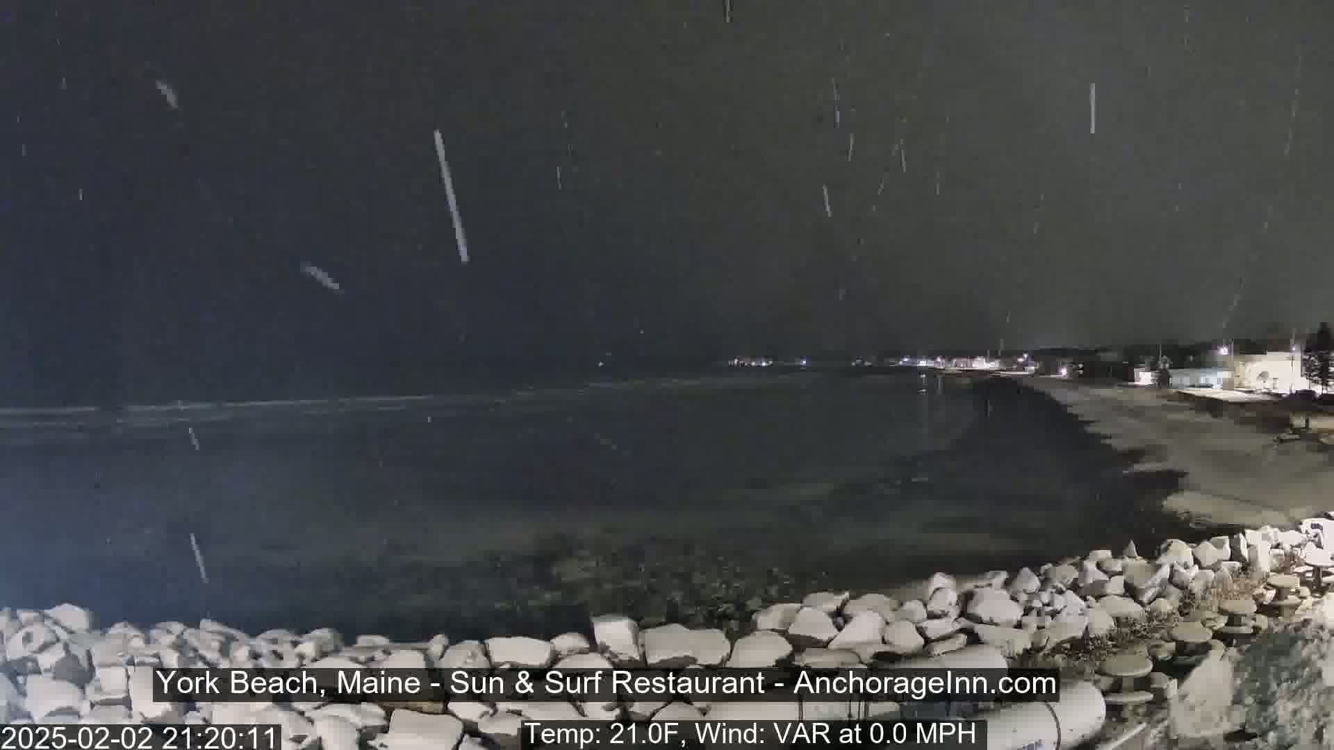 A nighttime beach scene shows light snow falling over a dark ocean and shoreline, with a rocky embankment in the foreground.