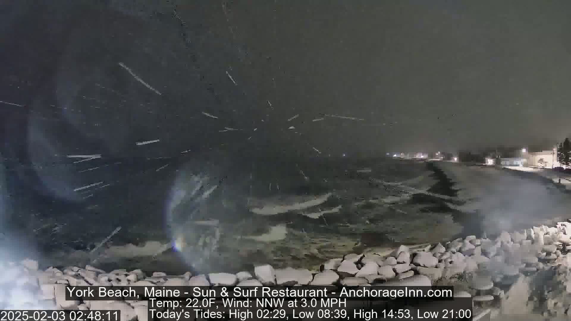 A nighttime view of a snow-covered rocky shoreline with rough waves crashing against it during a light snowfall.