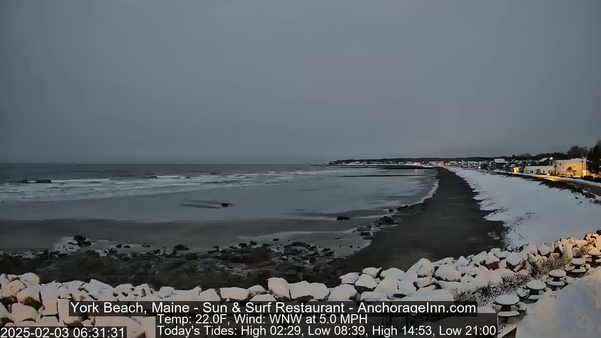 A snow-covered beach stretches along a calm, gray ocean under an overcast sky.