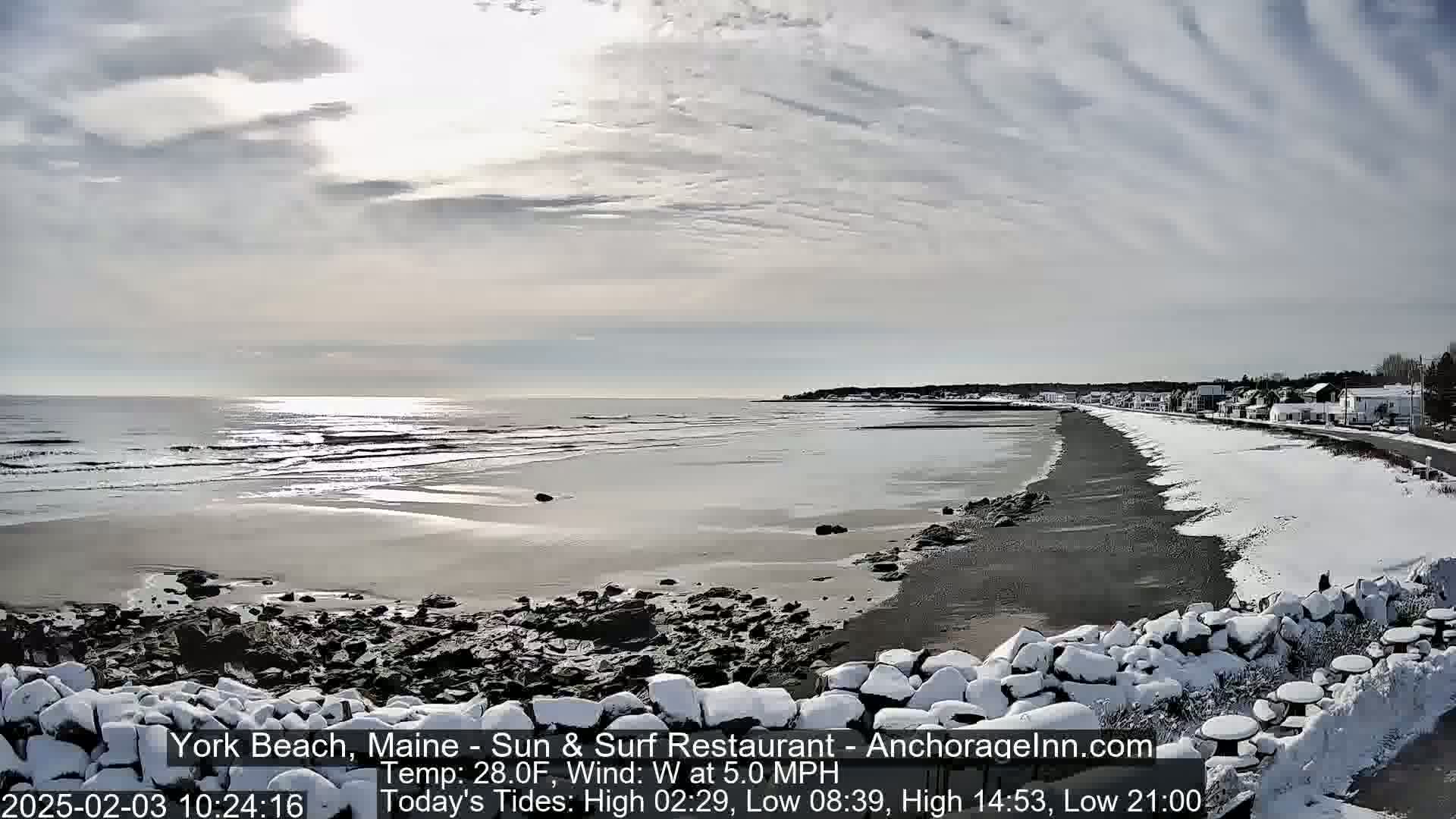 A snow-covered beach with a calm ocean and a town in the distance under a partly cloudy sky.