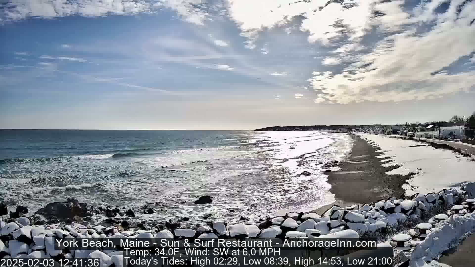 A snow-dusted beach with dark sand and waves rolling onto the shore under a partly cloudy sky.